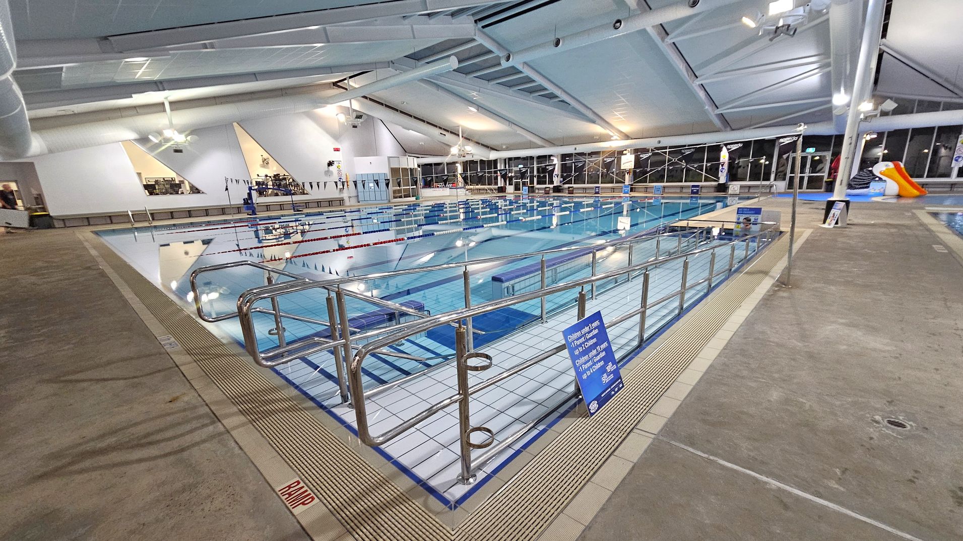 An indoor swimming pool with multiple lanes, surrounded by a metal railing. The pool area has a tiled edge and a ramp marked with "RAMP" in red. On the right, there's a small orange and white slide. The ceiling is high with exposed beams and lighting fixtures. Large windows line the right side, and there are triangular windows on the left wall. The floor is concrete, and there are signs with information around the pool.
