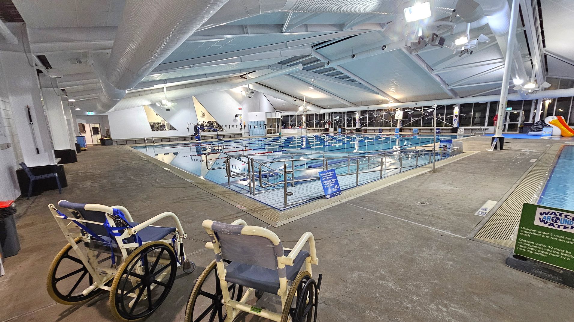 Indoor swimming pool area with two wheelchairs on the left side, positioned on a brown and green speckled floor. The pool is in the centre, surrounded by a metal railing. Overhead, a white ceiling with large ducts and lights. On the right, a slide and a sign with safety instructions. Large windows line the far wall, allowing natural light to enter.