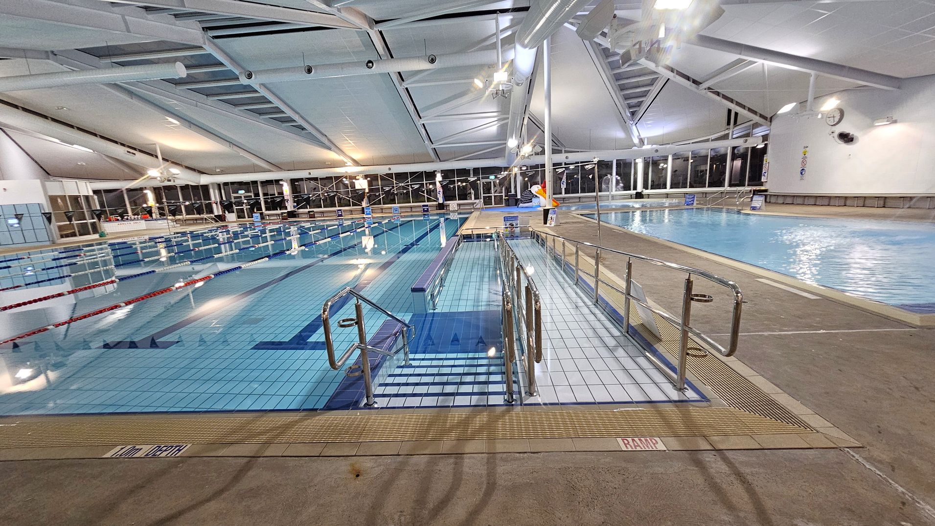 Indoor swimming pool area with a tiled floor. On the left, there are lanes marked with red and blue ropes. In the centre, a ramp with metal railings leads into the water. On the right, a smaller pool is visible. The ceiling is high with exposed beams and bright lighting. Large windows line the walls, and a clock is mounted on the far wall. The area is spacious and well-lit.