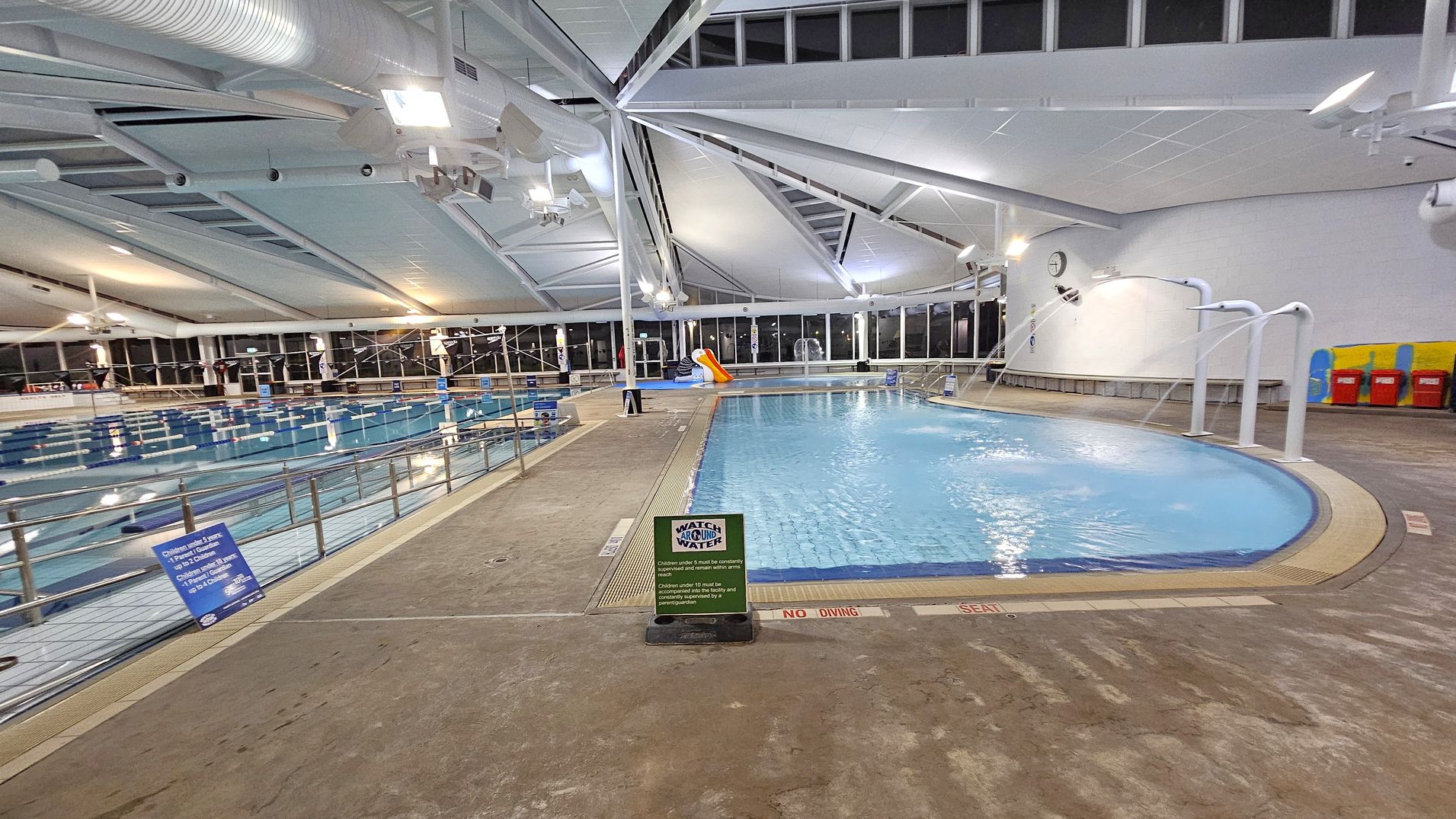 Indoor swimming pool area with two pools. On the left, a large lap pool with lane dividers and a railing along the edge. On the right, a smaller leisure pool with water features and a slide. The floor is textured concrete with safety signs. The ceiling is high with exposed beams and ductwork, and large windows line the walls, allowing natural light. A clock is mounted on the wall above benches and colourful lockers.