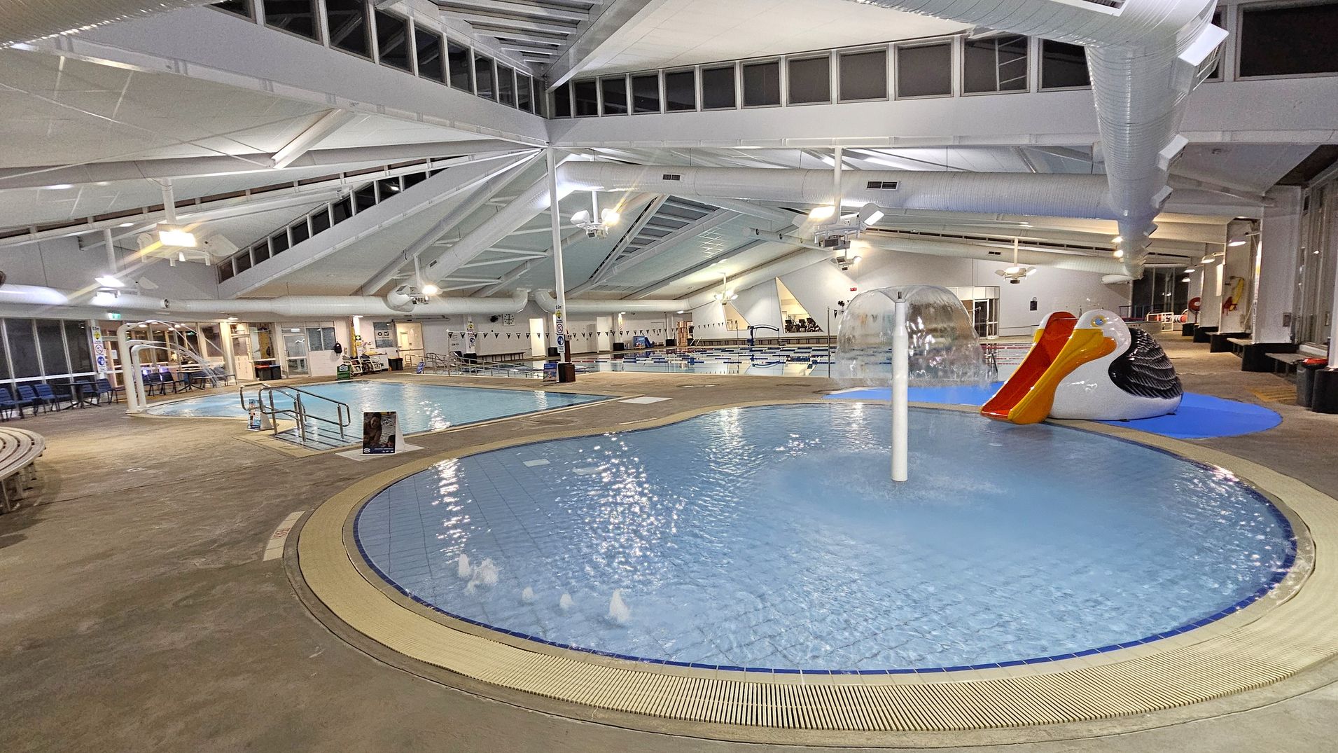 Indoor swimming pool area with a shallow, circular pool in the foreground featuring a water fountain. To the right, a large pelican-shaped slide with a red chute is visible. The background shows a larger pool with lanes and seating areas. The ceiling is high with exposed beams and ductwork, and the floor is a mix of tiles and concrete. Bright overhead lights illuminate the space, and large windows line the walls, providing natural light.
