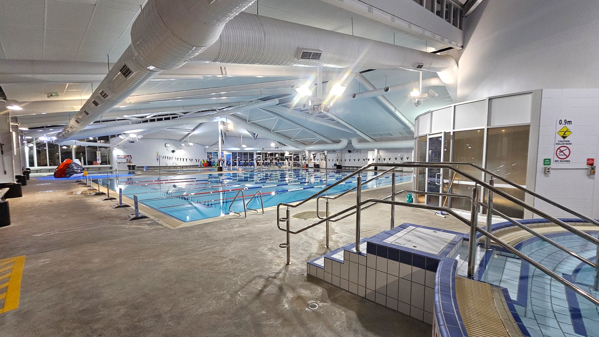 An indoor swimming pool area with a tiled floor. On the right, there are metal railings leading into a small pool section. The main pool is in the centre, with lane dividers and a depth sign indicating 0.9 metres. Large windows and a clock are visible in the background. Overhead, there are large white ventilation ducts and a high ceiling with lights. On the left, there is a colourful inflatable structure near the poolside.