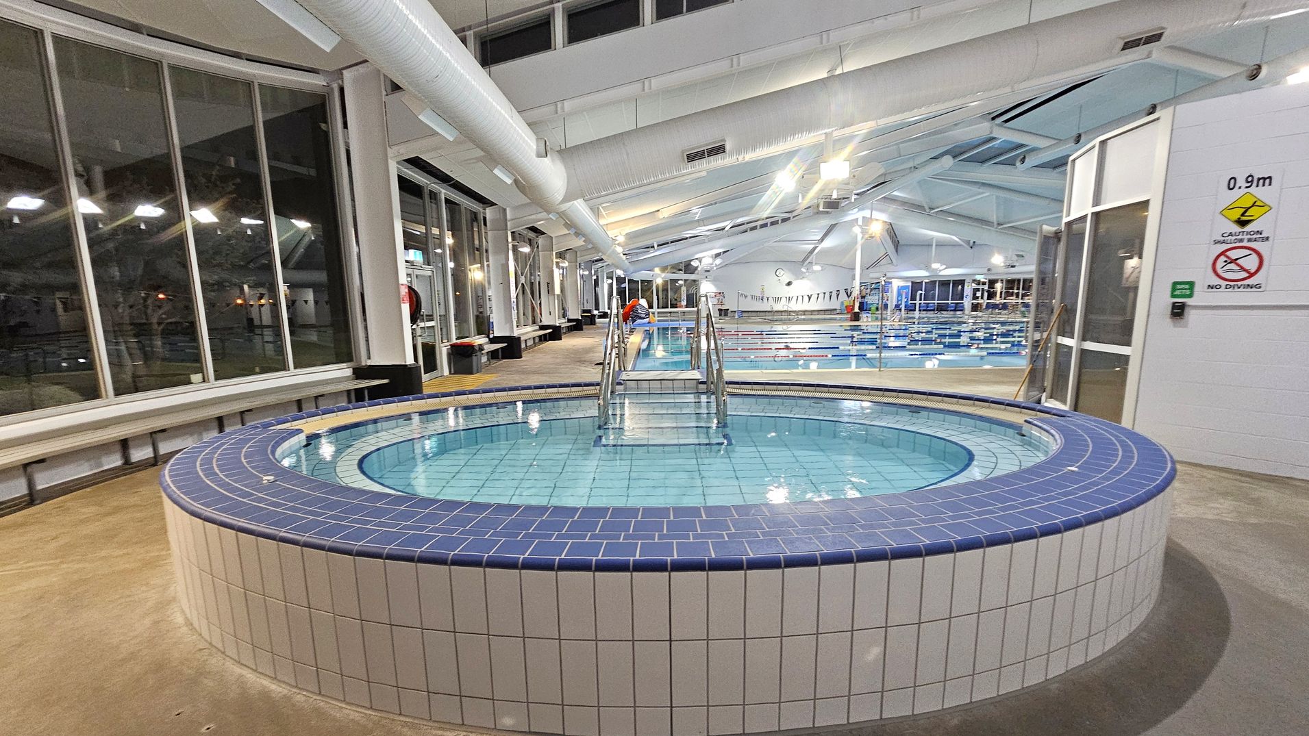 A spacious indoor swimming facility with a round, tiled hot tub in the foreground, featuring blue and white tiles. A metal handrail leads into the water. To the right, a sign indicates a depth of 0.9 metres and warns against diving. In the background, a larger swimming pool with lanes is visible, along with a high ceiling featuring exposed ducts and bright lighting. Large windows line the left side, with benches along the wall.