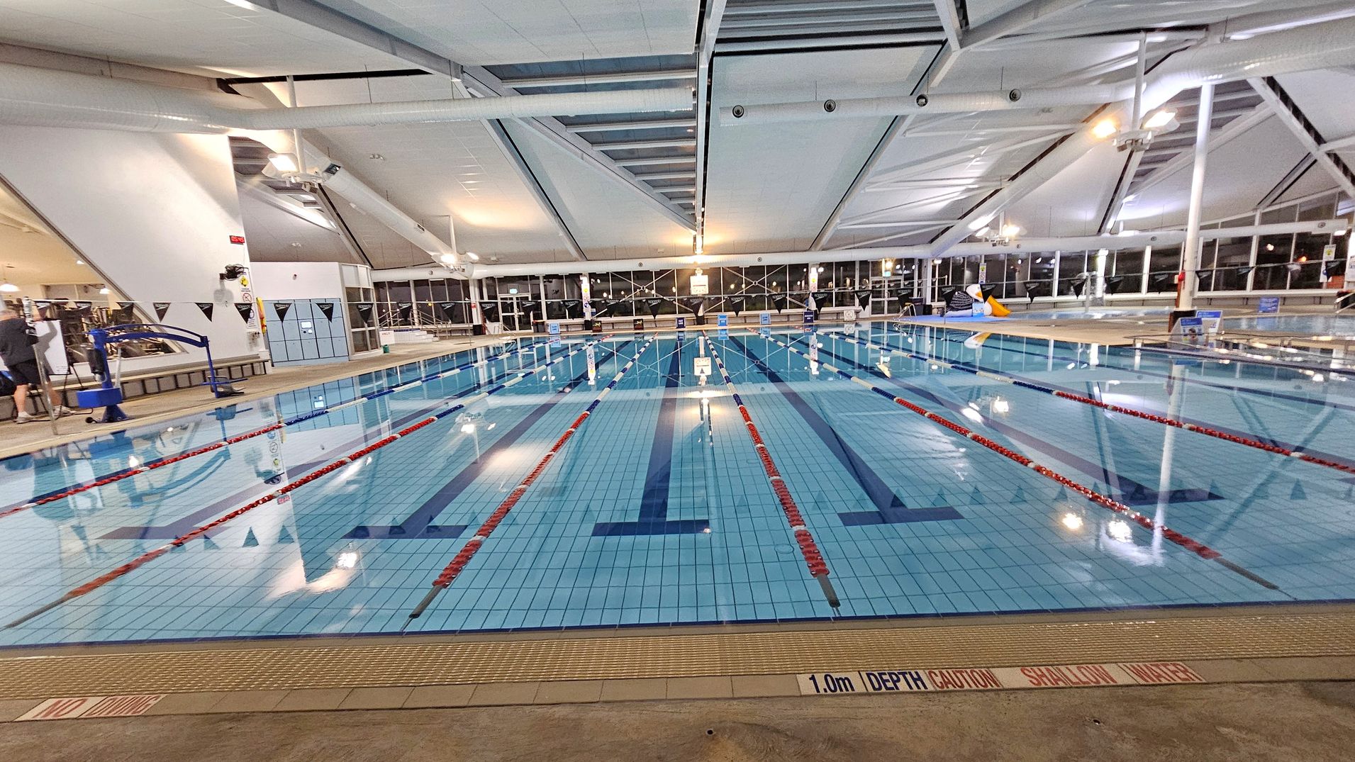 An indoor swimming pool with multiple lanes marked by red and white lane dividers. The pool's floor is tiled in light blue with dark blue lane markings. On the left, there is a pool lift and a person standing near the edge. The right side features a large inflatable penguin. The ceiling is high with exposed beams and lighting fixtures. Large windows line the back wall, allowing natural light to enter. The pool deck is tiled, and a sign indicates a depth of 1.0m.