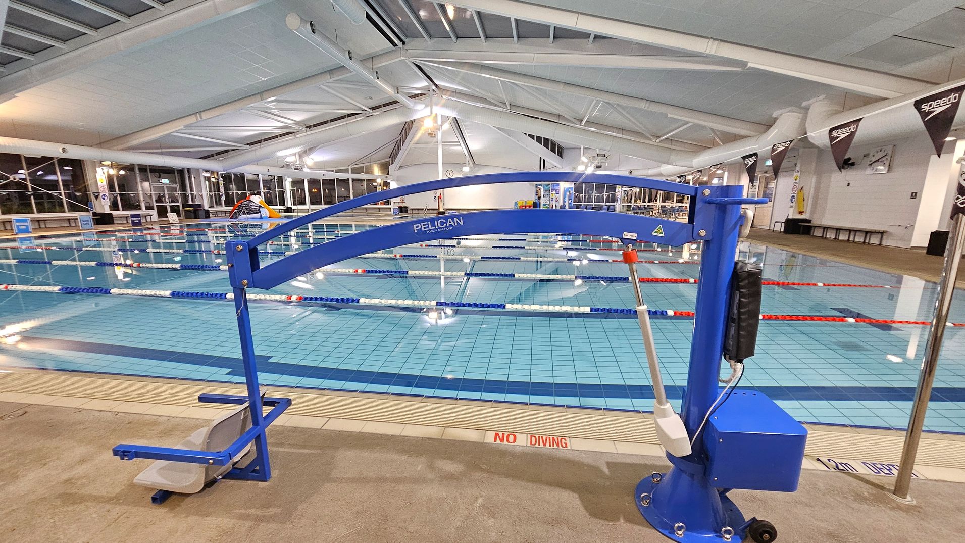An indoor swimming pool with multiple lanes separated by floating lane dividers. In the foreground, a blue pool lift labelled "Pelican" is positioned on the pool deck, designed for accessibility. The pool area is well-lit with large windows on the left side and a high, white ceiling with exposed beams. The floor is tiled, and there are "No Diving" warnings near the pool edge. Banners with the "Speedo" logo hang from the ceiling on the right side.