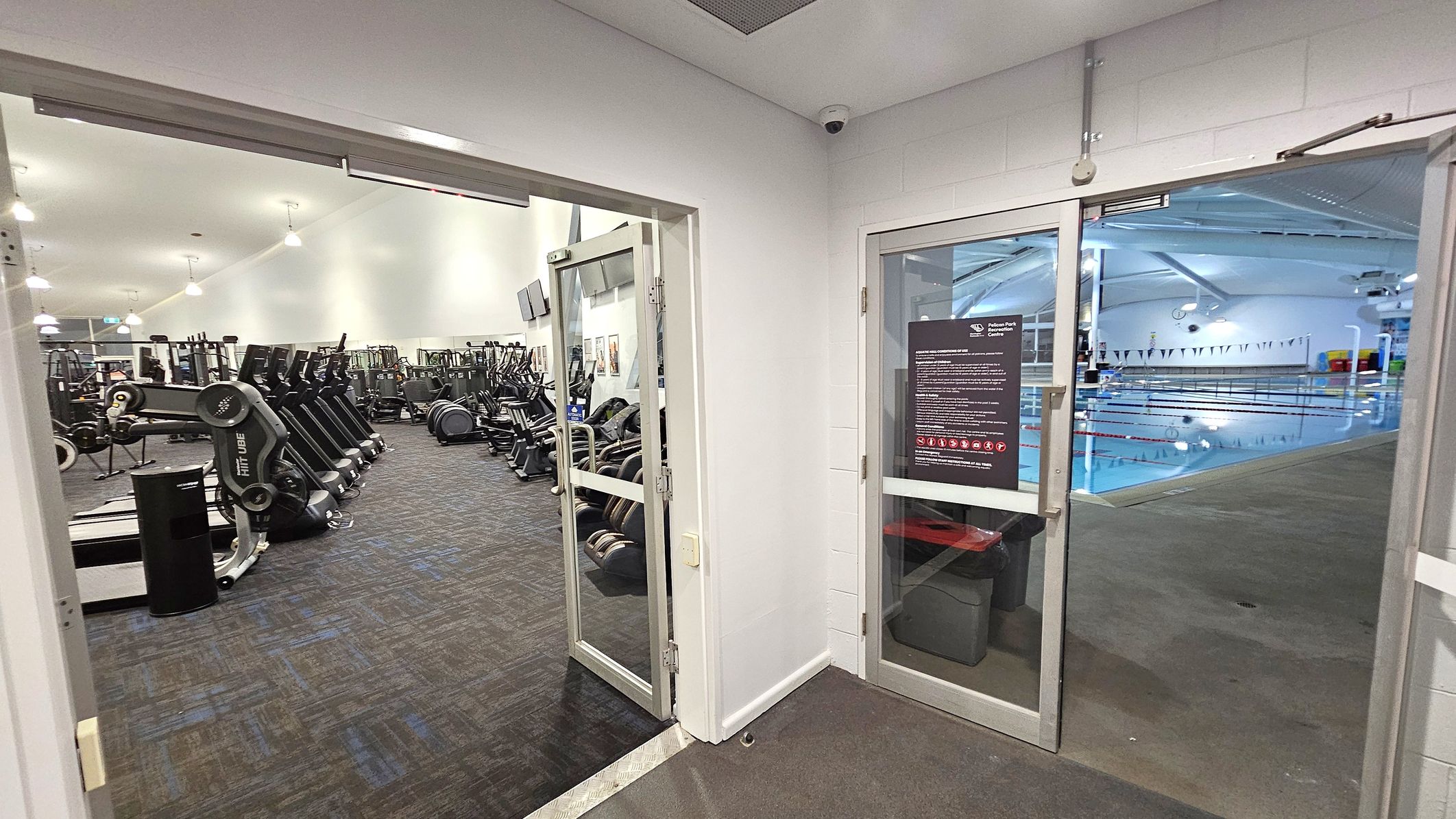 On the left, there is a gym area with various exercise equipment, including treadmills and stationary bikes, arranged in rows on a carpeted floor. On the right, there is a swimming pool with lanes marked by floating dividers, visible through a glass door. The pool area has a tiled floor and a high ceiling with lights. The walls are white, and there is a security camera mounted in the corner above the doors.