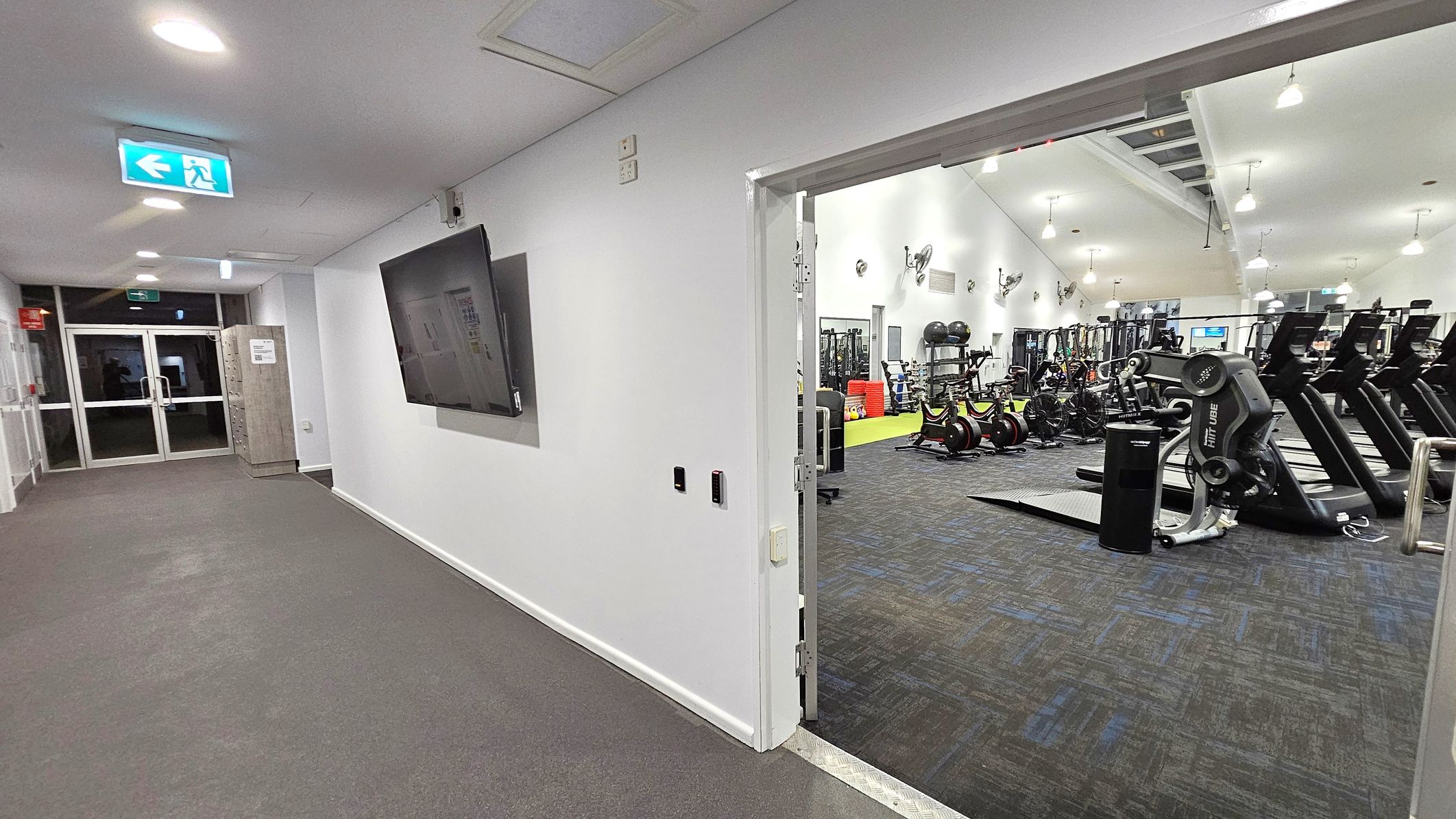A gym entrance with a hallway on the left featuring a grey carpet and a wall-mounted screen. An exit sign is visible on the ceiling. At the end of the hallway, there are glass double doors and a wooden cabinet. On the right, an open door leads into a gym area with various exercise equipment, including treadmills and stationary bikes. The gym has a high ceiling with hanging lights and a dark carpeted floor.