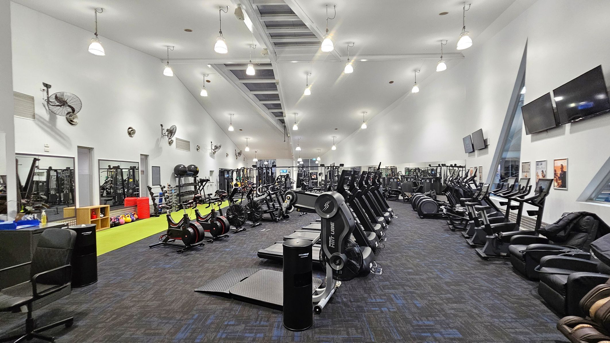 A spacious gym interior with a high ceiling and bright lighting. On the left, there are wall-mounted fans, mirrors, and a variety of gym equipment including weights and exercise balls. In the centre, rows of exercise machines such as treadmills and stationary bikes are neatly arranged. On the right, there are large windows, wall-mounted televisions, and more exercise machines. The floor is carpeted in dark tones, and the ceiling features hanging lights.