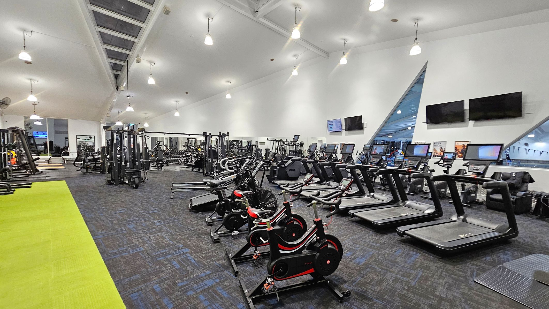 A spacious gym with a high, sloped ceiling and bright lighting. On the right, there are several treadmills and exercise bikes lined up, with two large flat-screen TVs mounted on the wall above. The left side features various weight machines and a green exercise mat area. The floor is covered with dark patterned carpet. Triangular windows on the right wall provide a view into another area. The ceiling has multiple hanging lights.