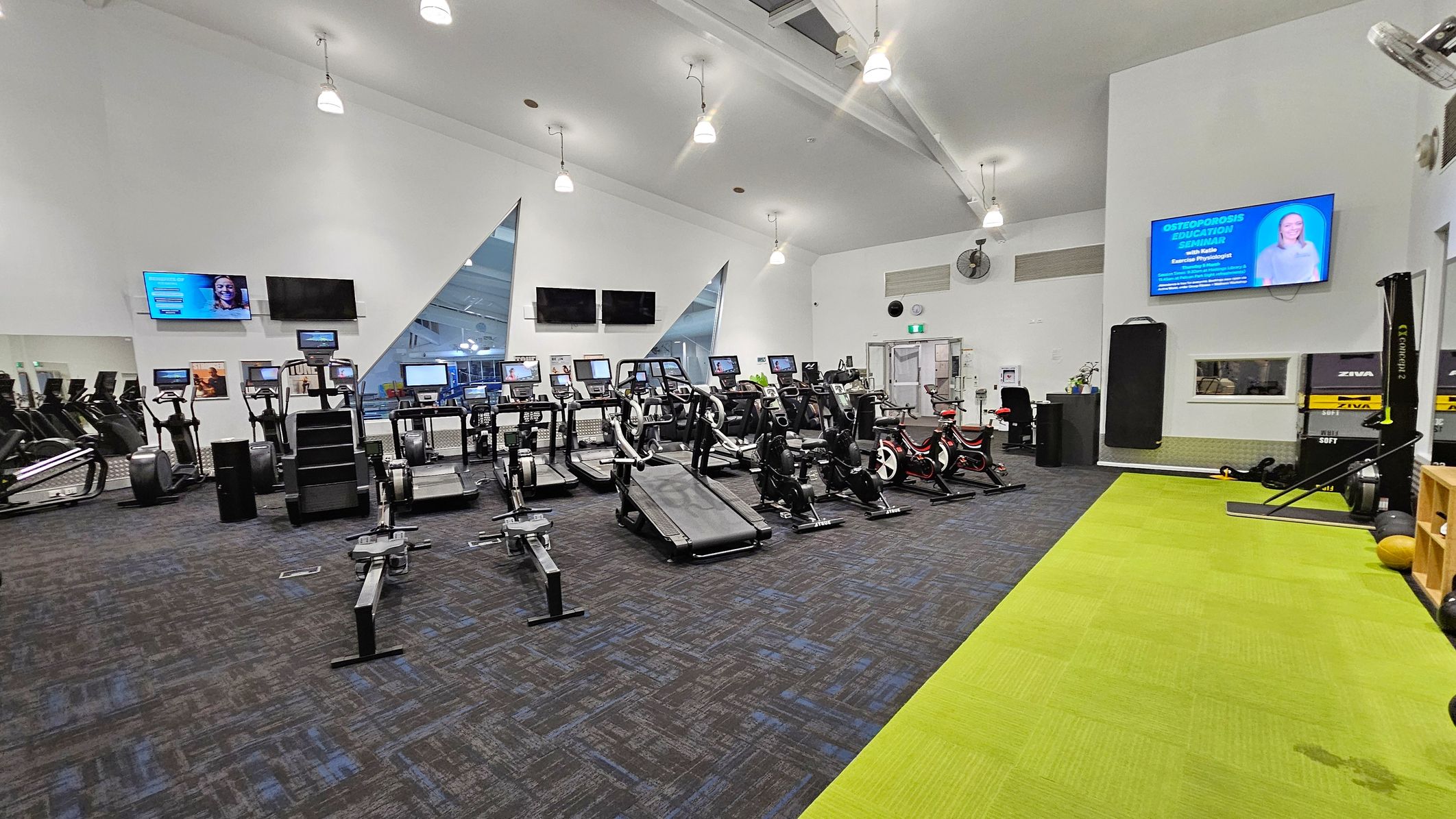 A spacious gym with a variety of exercise equipment. On the left, there are multiple treadmills and elliptical machines facing a wall with three mounted screens. In the centre, rowing machines and a treadmill are arranged neatly. On the right, stationary bikes and a rowing machine are visible. The floor is a mix of dark and bright green carpet. The ceiling is high with hanging lights, and a large screen is mounted on the right wall displaying information.