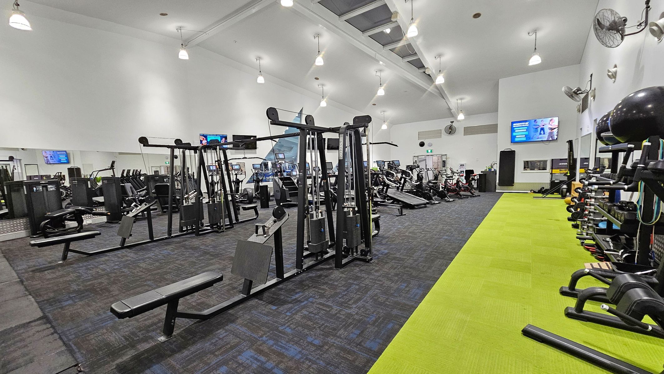 A spacious gym interior with a variety of exercise equipment. On the left, there are weight machines and cardio equipment like treadmills and ellipticals. In the centre, more weight machines are arranged in rows. On the right, a bright green mat runs along the floor with free weights and exercise balls on racks. The ceiling is high with multiple hanging lights, and there are several wall-mounted fans and televisions. The floor is a mix of dark carpet and green matting.