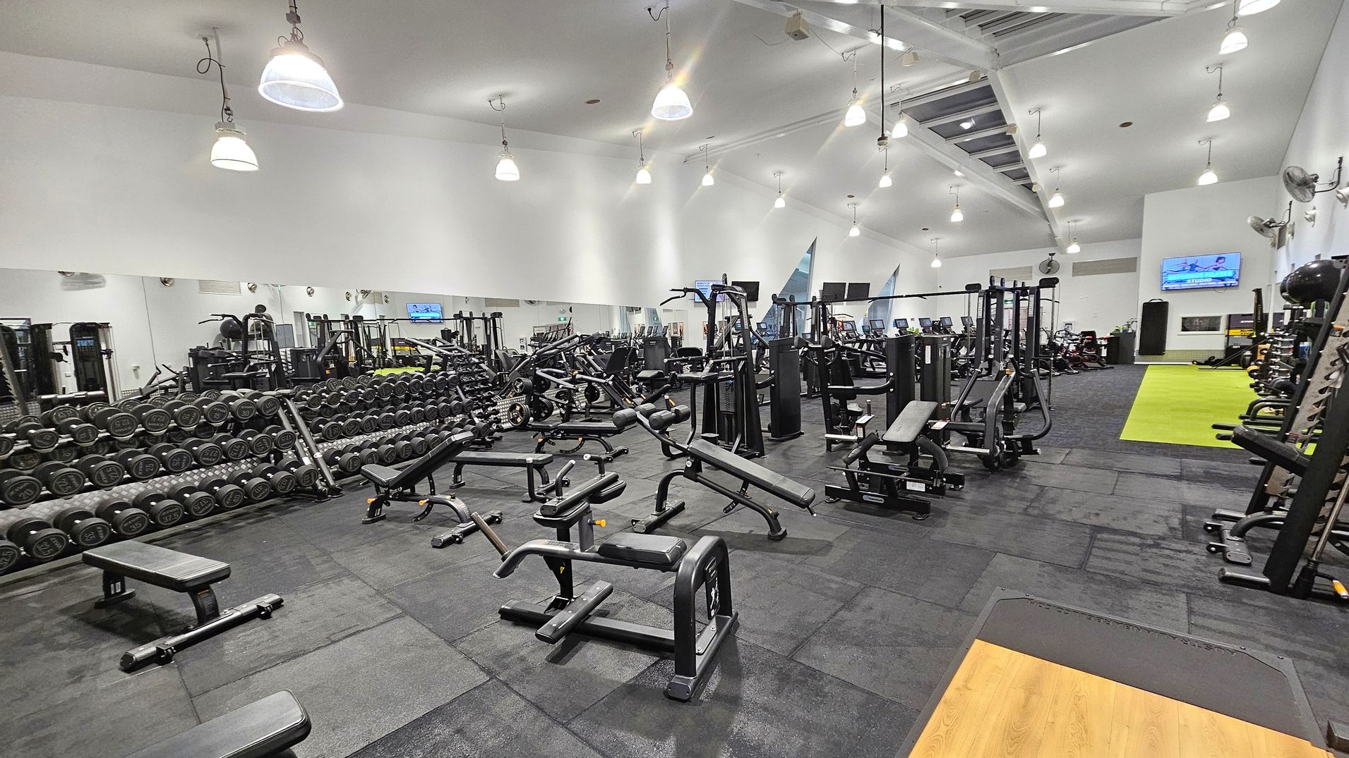 A spacious gym interior with a variety of exercise equipment. On the left, there are rows of dumbbells neatly arranged on racks in front of a large mirror. In the centre, various weight machines and benches are scattered across the black rubber flooring. On the right, there is a green turf area and more exercise machines. The ceiling is high with exposed beams and multiple hanging lights. A television is mounted on the far wall.