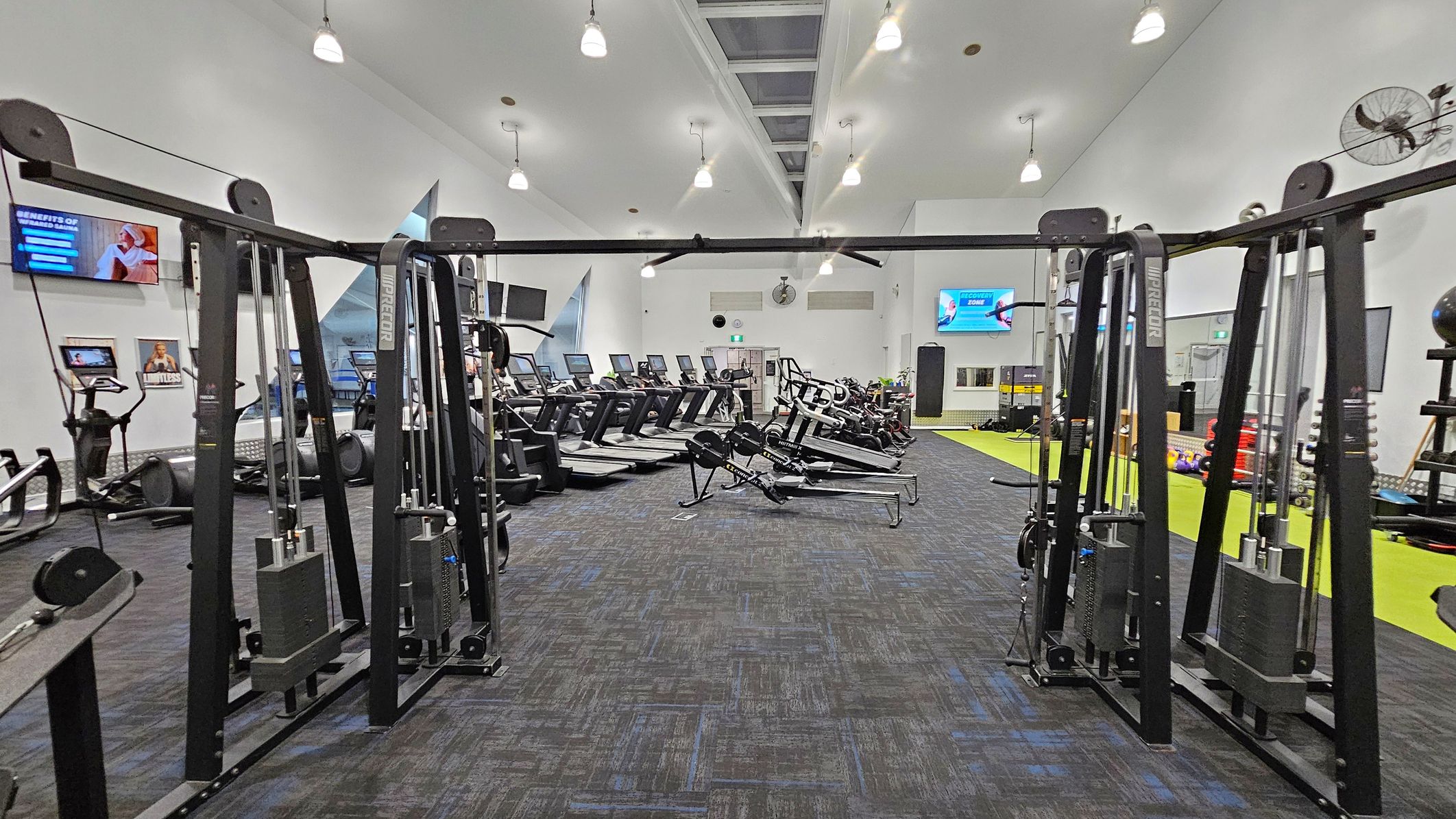 A spacious gym interior with a variety of exercise equipment. In the foreground, there are weight machines with cables and pulleys. The middle area features several treadmills and rowing machines lined up. On the left wall, there are mounted screens and posters. The right side has a bright green mat area with additional equipment like dumbbells. The ceiling is high with multiple hanging lights, and the floor is carpeted in dark grey.