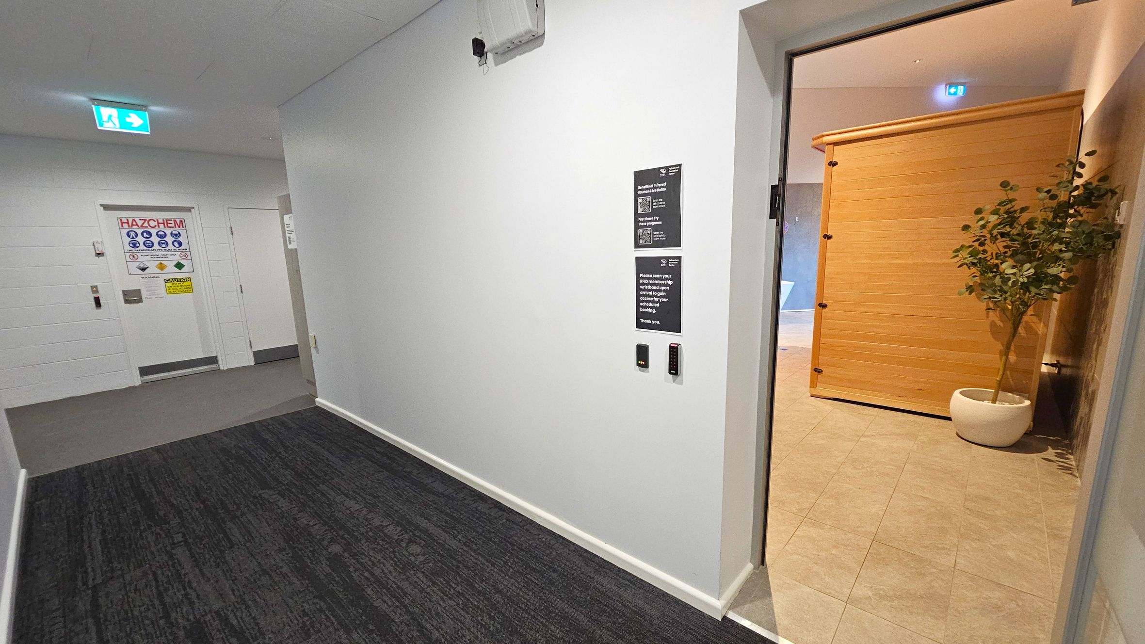 A corridor with a dark carpeted floor and white walls. On the left, a door marked "HAZCHEM" with various safety signs. Above it, an illuminated green exit sign. On the right, an open door leads to a room with a light-coloured tiled floor, a wooden structure, and a potted plant. The wall near the open door has signs and a card reader. The ceiling is plain with a few fixtures.