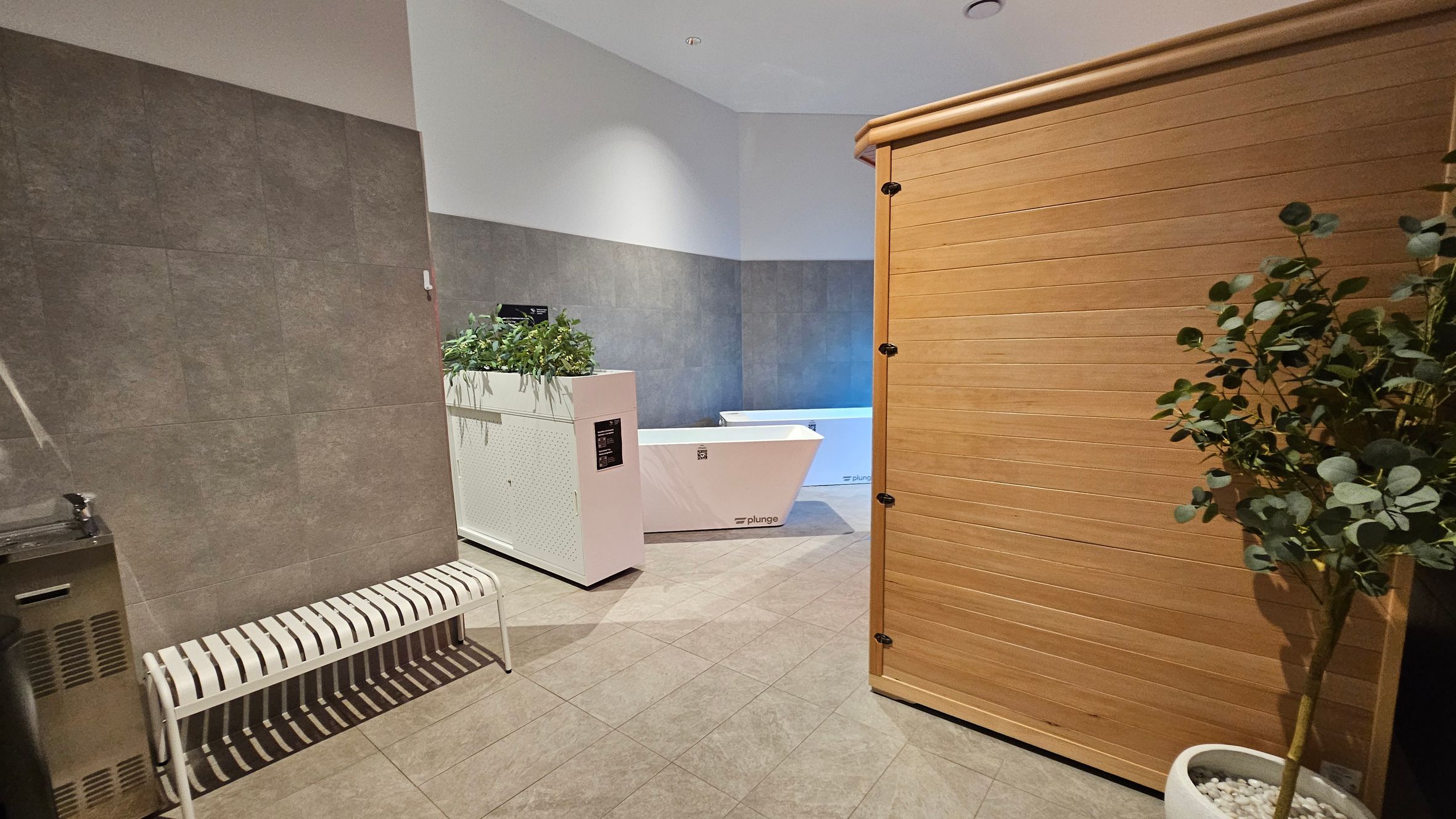 A room with a tiled floor and a light-coloured ceiling. On the left, there's a metal bench and a water fountain. In the centre, a white plunge tub is partially visible, with a planter box containing greenery on top. On the right, a wooden structure, possibly a sauna, is present. A potted plant with green leaves is in the foreground on the right side. The walls are a mix of grey and white tones.