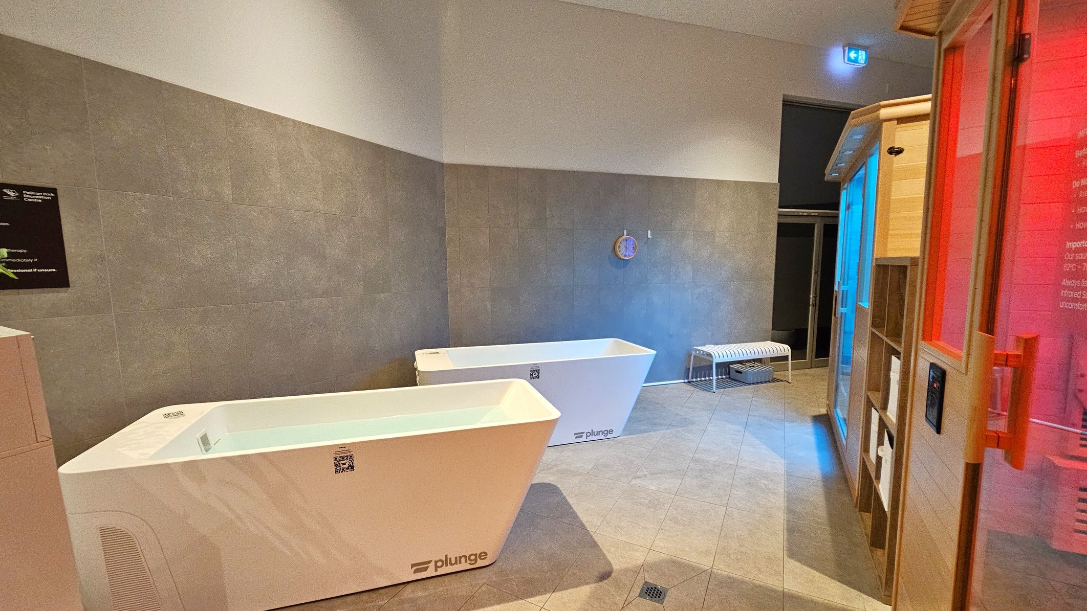On the left, there are two white plunge tubs with a modern design, placed against a grey-tiled wall. In the middle, a small clock is mounted on the wall above a white bench. On the right, a wooden sauna with a glass door emits a red glow, and a shelf unit is visible beside it. The floor is tiled in a light colour, and the ceiling is plain with a blue exit sign above the door in the background.
