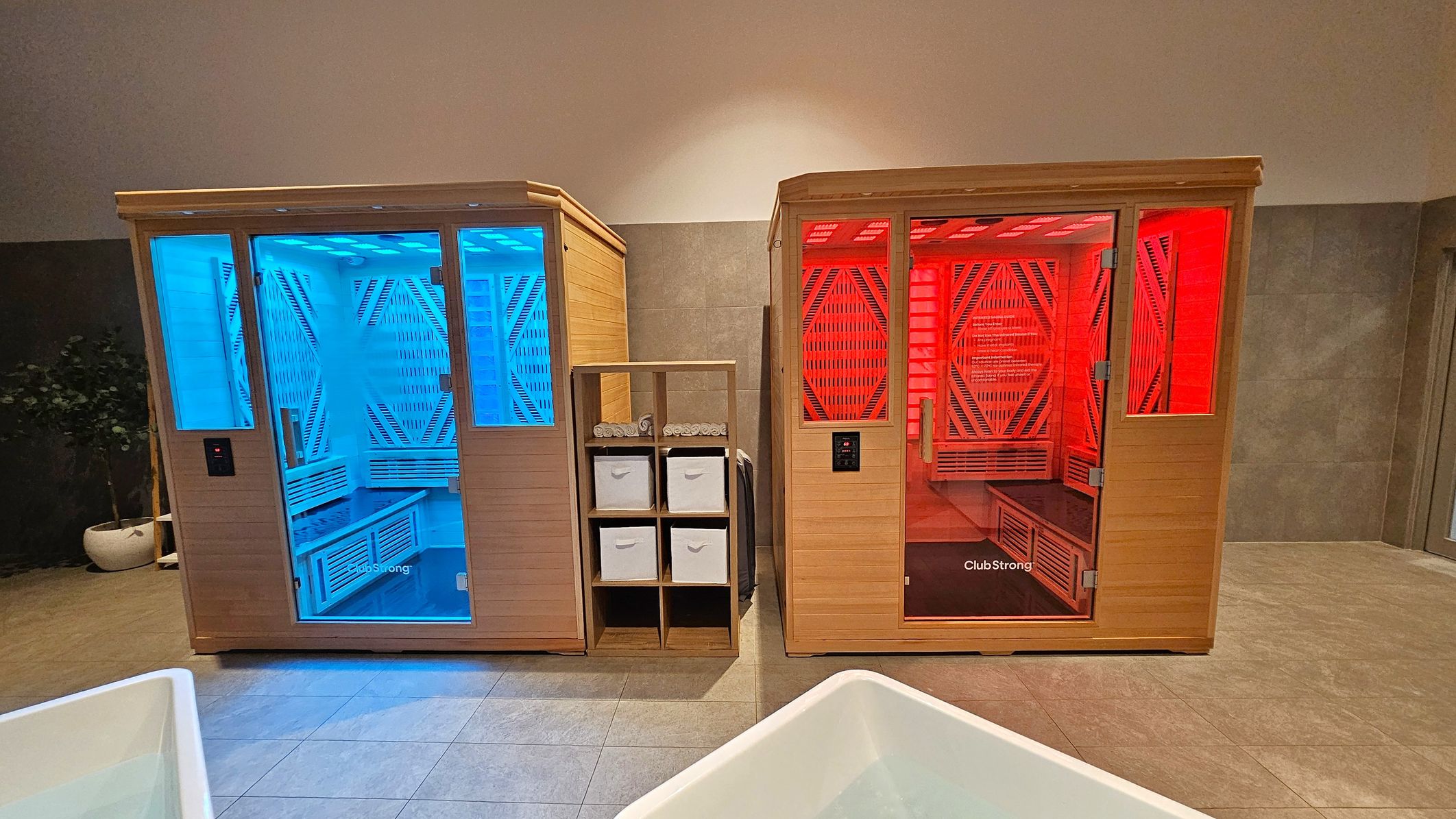 Two wooden infrared saunas are side by side. The left sauna glows with blue light, while the right one emits red light. Between them is a wooden shelving unit with rolled white towels and storage boxes. The floor is tiled in a light grey, and the background wall is a mix of grey and white. A potted plant is partially visible on the far left.