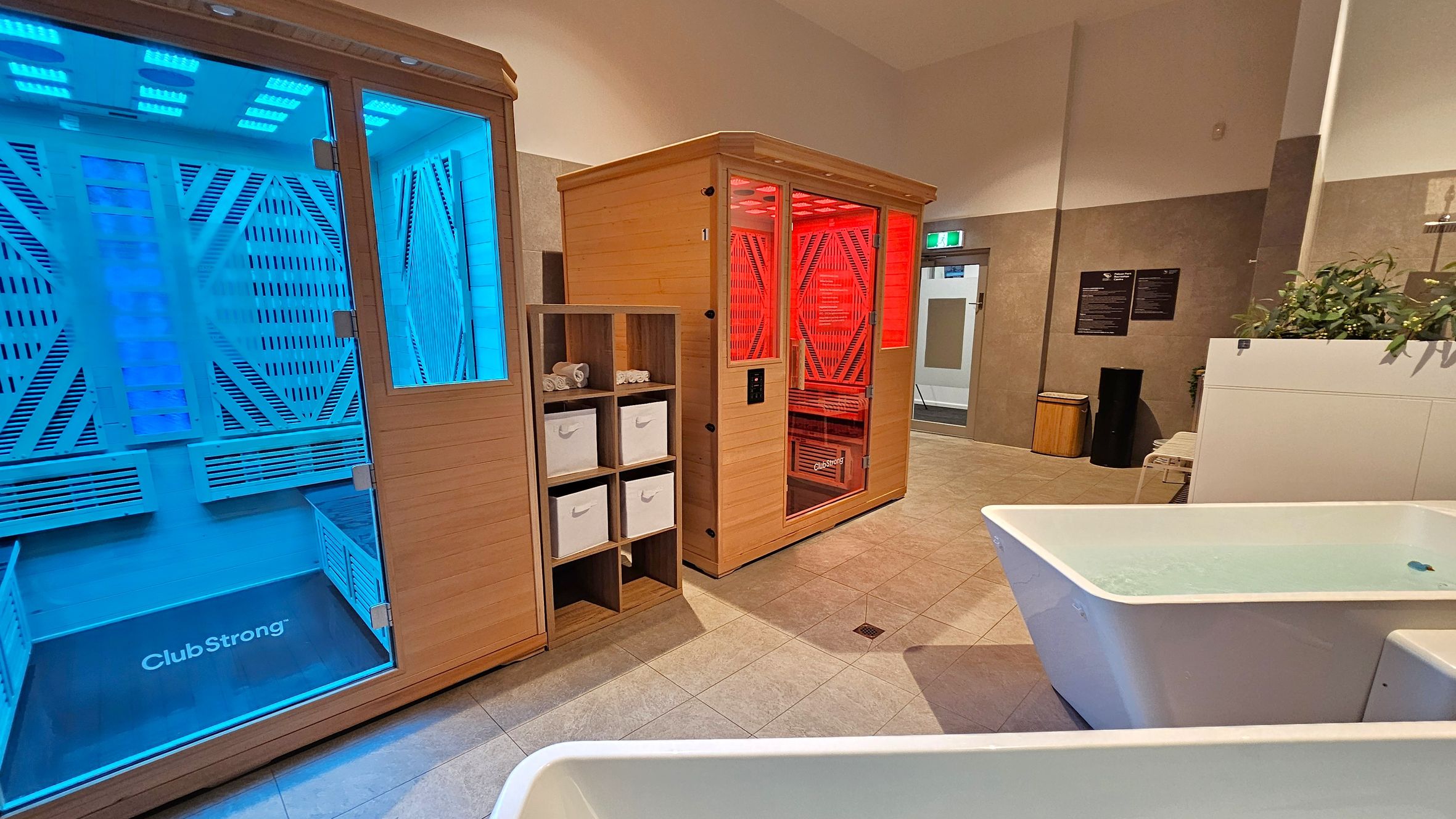 On the left, a wooden sauna with blue lighting inside. In the centre, another wooden sauna with red lighting. Between them, a wooden shelving unit with white storage boxes and rolled towels. On the right, a white bathtub filled with water, and a plant on a counter. The floor is tiled, and the ceiling is curved. In the background, a door with an exit sign, a waste bin, and wall-mounted signs.