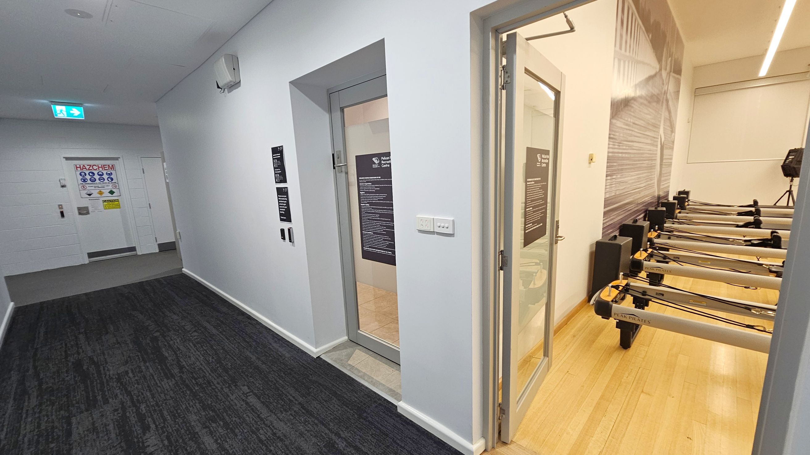 A hallway with a dark carpeted floor and white walls. On the left, a door marked "HAZCHEM" with safety signs. On the right, an open door leads to a room with several Pilates reformer machines on a light wooden floor. The room has a large wall graphic and a window with a blind. The ceiling has recessed lighting and an emergency exit sign is visible above the hallway.