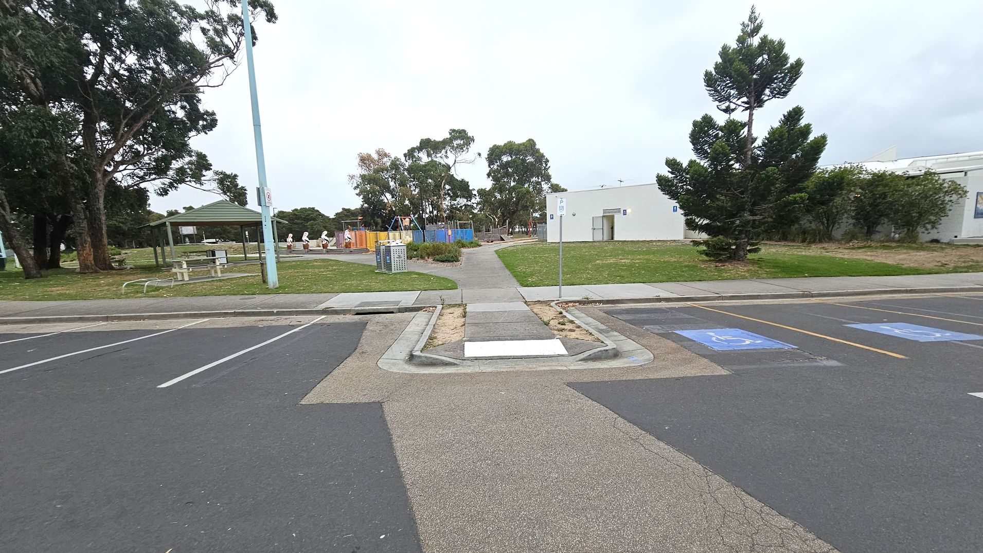A paved pathway leads from the foreground across a parking area with marked disabled parking spaces. On the left, there are large trees and a sheltered picnic area with tables. In the centre, a playground with colourful equipment is visible, surrounded by more trees. On the right, a white building with a tree in front is situated on a grassy area. The scene is open and spacious, with a mix of natural and built elements.