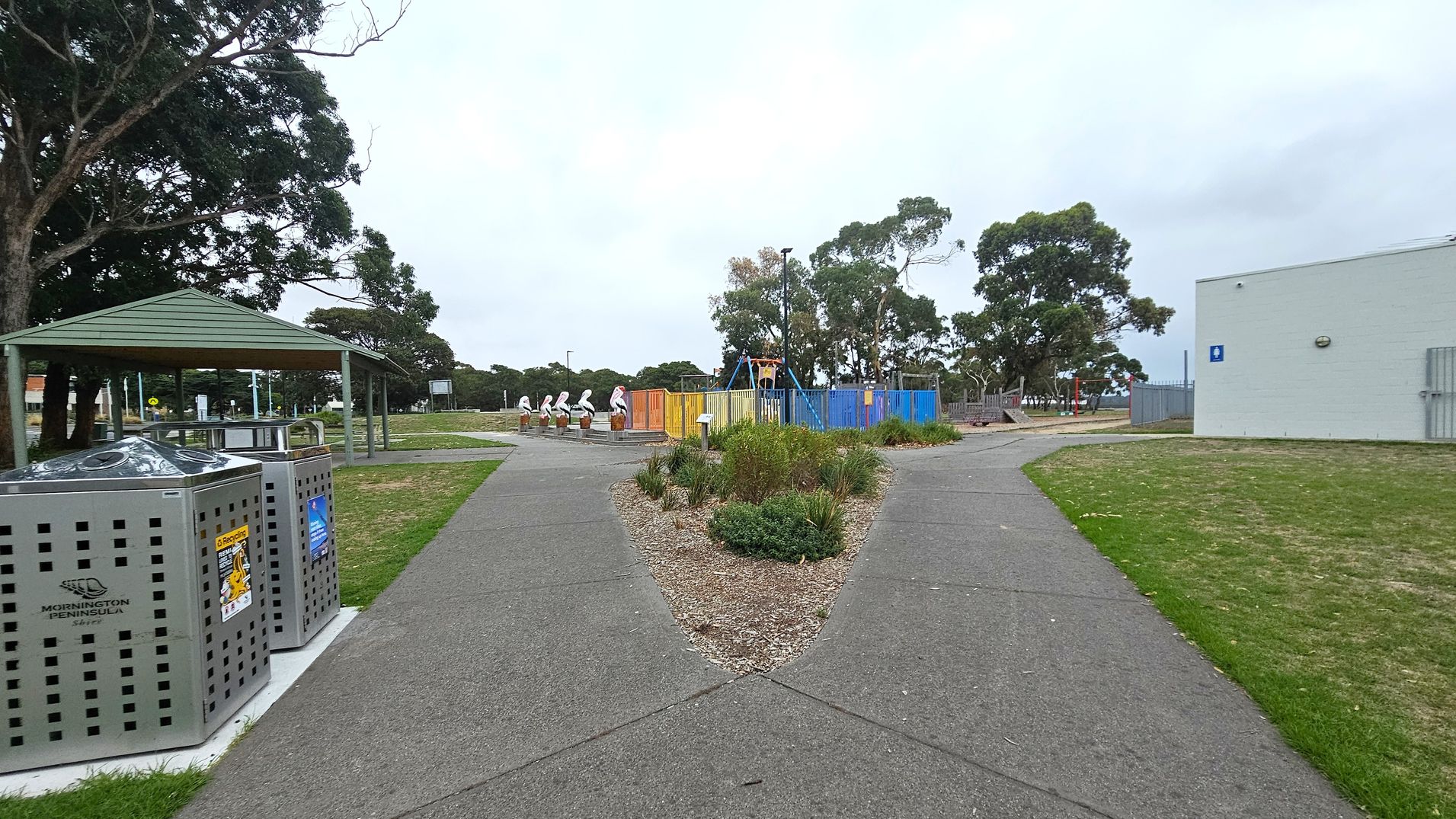A park scene with a paved path splitting into two directions. On the left, there is a sheltered picnic area with metal bins labelled "Mornington Peninsula Shire" nearby. In the centre, a colourful fenced playground with swings and pelican sculptures is visible. On the right, a light-coloured building with a blue sign is adjacent to a grassy area. Trees are scattered throughout the background.