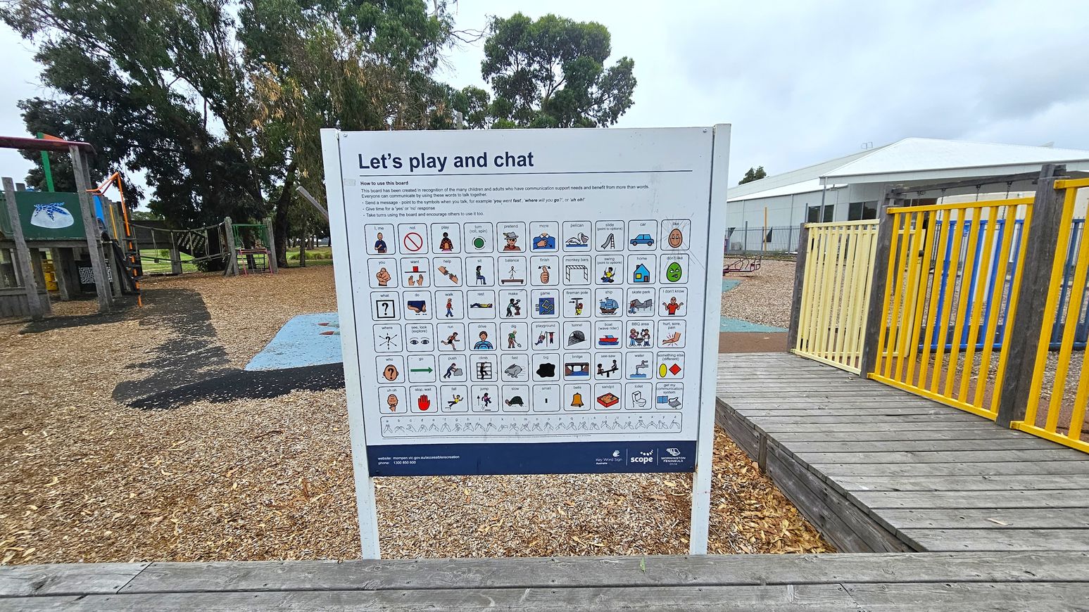 A communication board is in the centre, featuring various symbols and words for interaction. To the left, there's a playground with wooden structures and slides, surrounded by trees. On the right, a wooden walkway with yellow railings leads to more playground equipment. The ground is covered with wood chips, and a building with a white roof is visible in the background.