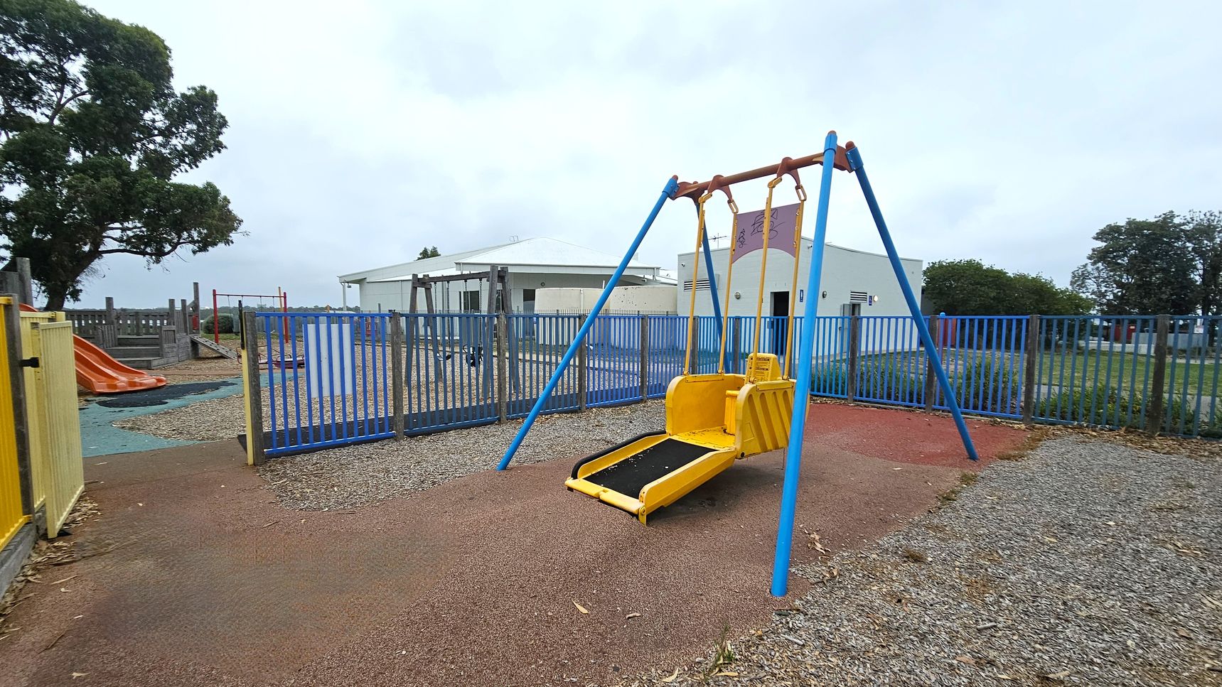 A playground with a yellow and blue swing set in the centre, designed for accessibility. To the left, there's an orange slide and a wooden play structure. The ground is covered with a mix of rubber and gravel. A blue metal fence encloses the area, with a yellow gate on the left. In the background, there are white buildings and trees.