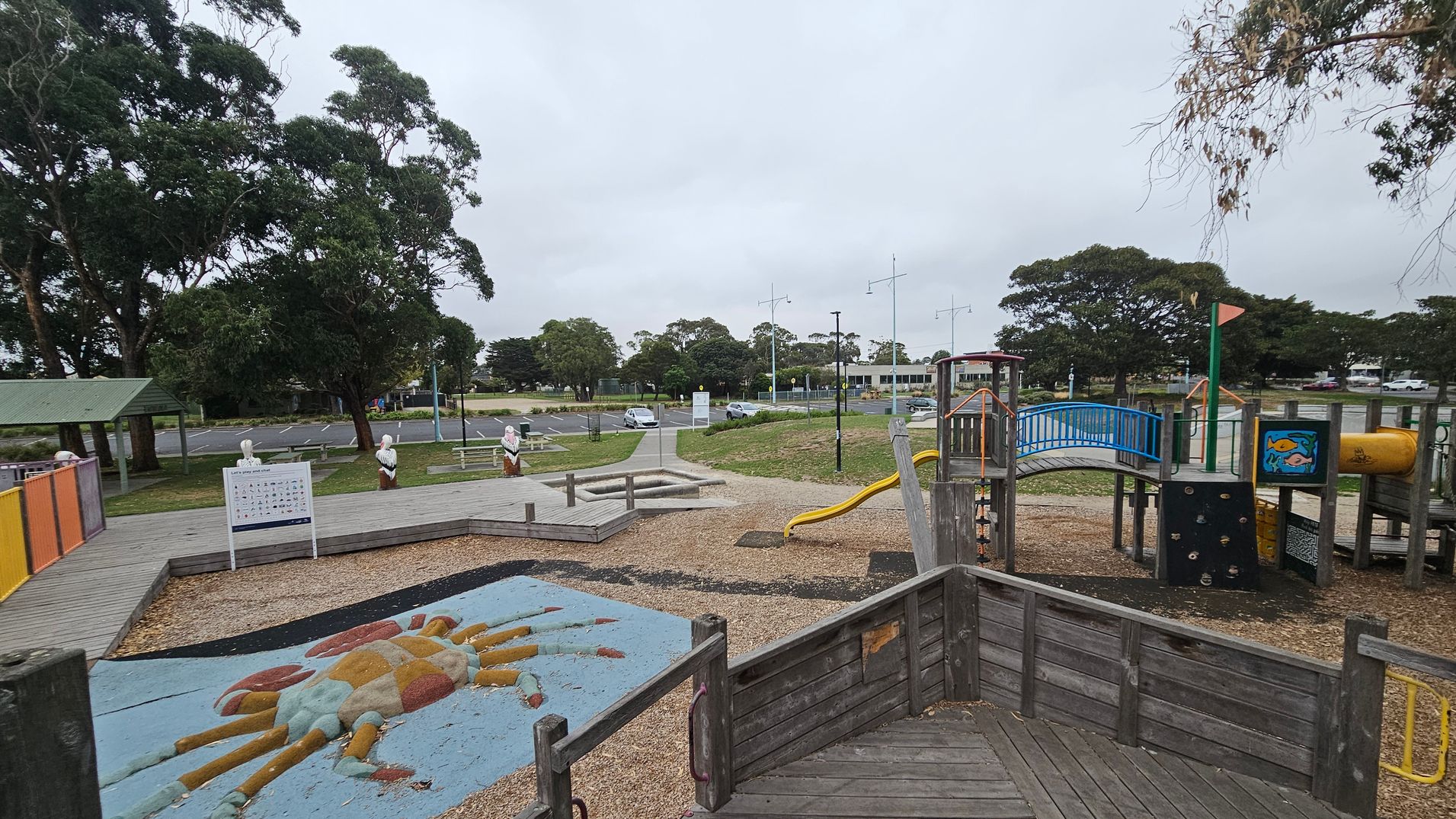 A playground with a wooden structure featuring a slide and climbing elements on the right. In the centre, a colourful crab design is embedded in the ground. To the left, there is a covered seating area with picnic tables and a signboard. The background shows a car park with several vehicles and trees lining the area. The ground is covered with wood chips, and there are pathways connecting different sections of the playground.