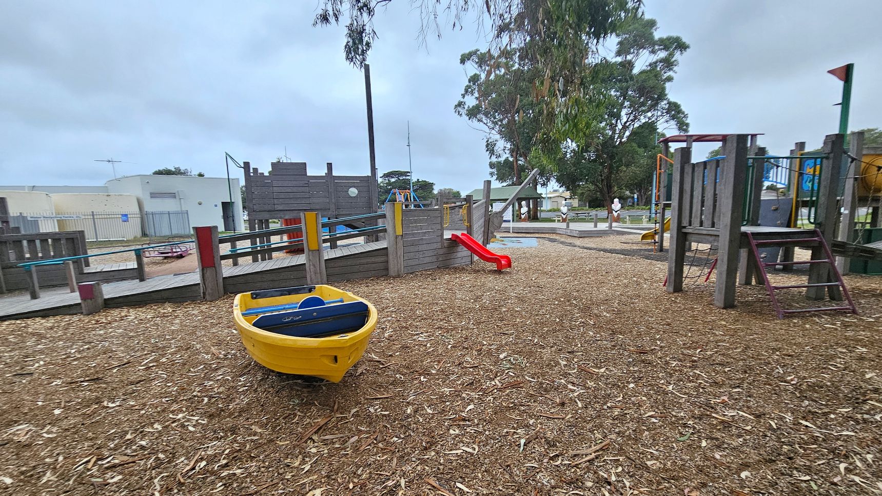 A playground with a woodchip-covered ground. On the left, a yellow play boat is visible. In the centre, a wooden play structure resembling a ship features a small red slide. To the right, more playground equipment is present, including platforms and climbing areas. In the background, there are trees and a white building with a fence.