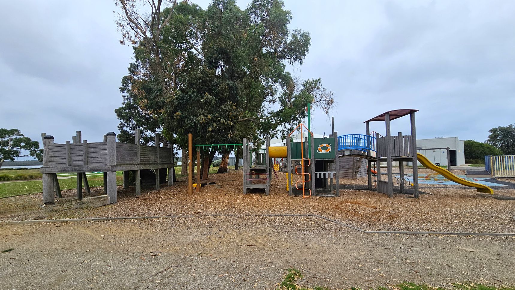 A playground with various equipment. On the left, there's a wooden structure resembling a fort. In the centre, a climbing frame with green monkey bars and a small slide. To the right, a larger structure with a blue bridge and a yellow slide. The ground is covered with wood chips and grass. In the background, there are trees and a building with a mural. The sky is overcast.