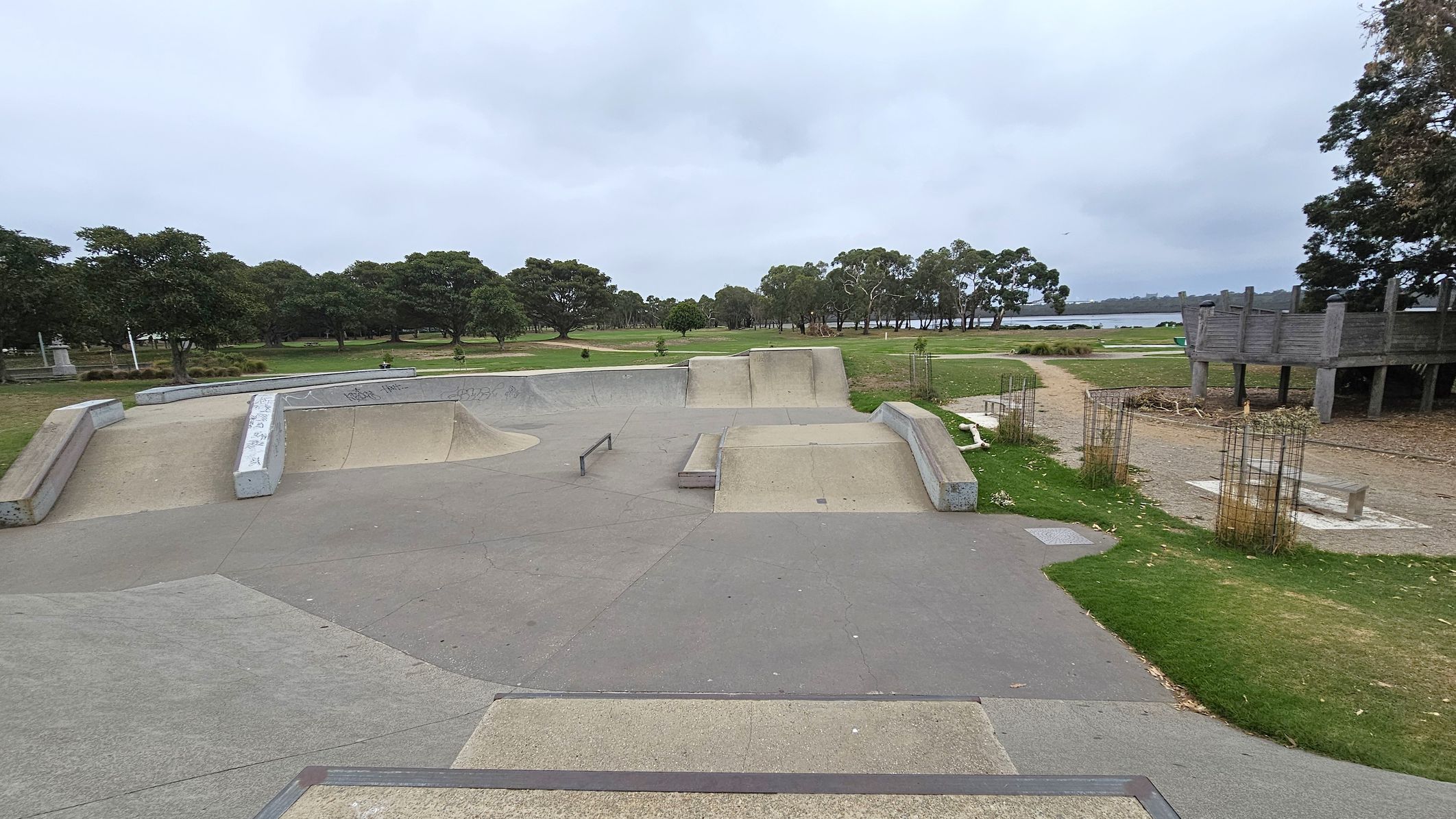 A skate park with concrete ramps and rails in the foreground. The ground is a mix of grey and green surfaces. To the right, there is a wooden structure on stilts and some grassy areas with young trees protected by wire cages. In the background, there are more trees and a body of water visible. The area is surrounded by greenery, with trees lining the horizon.