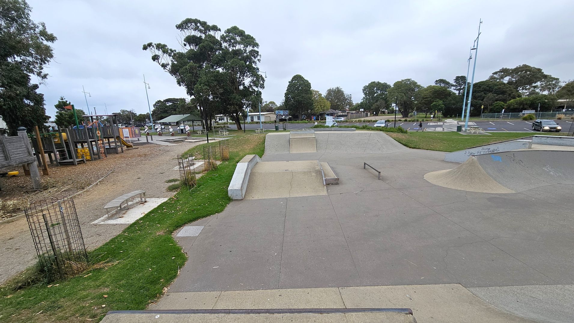 A skate park with concrete ramps and rails is in the foreground. To the left, there's a playground with wooden structures and slides. The background features a grassy area with trees and a few picnic tables. A car park with several vehicles is visible on the right. The ground is a mix of concrete and grass, and there are tall light poles scattered throughout the area.