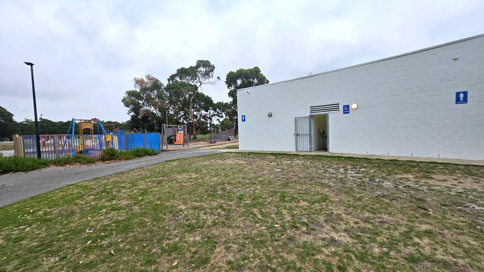 A grassy area with patches of dry grass in the foreground. On the right, a white building with a public toilet sign and an open entrance. On the left, a playground with blue fencing, swings, and climbing structures. Trees are visible in the background, and a pathway runs through the scene. A tall lamp post stands near the playground.