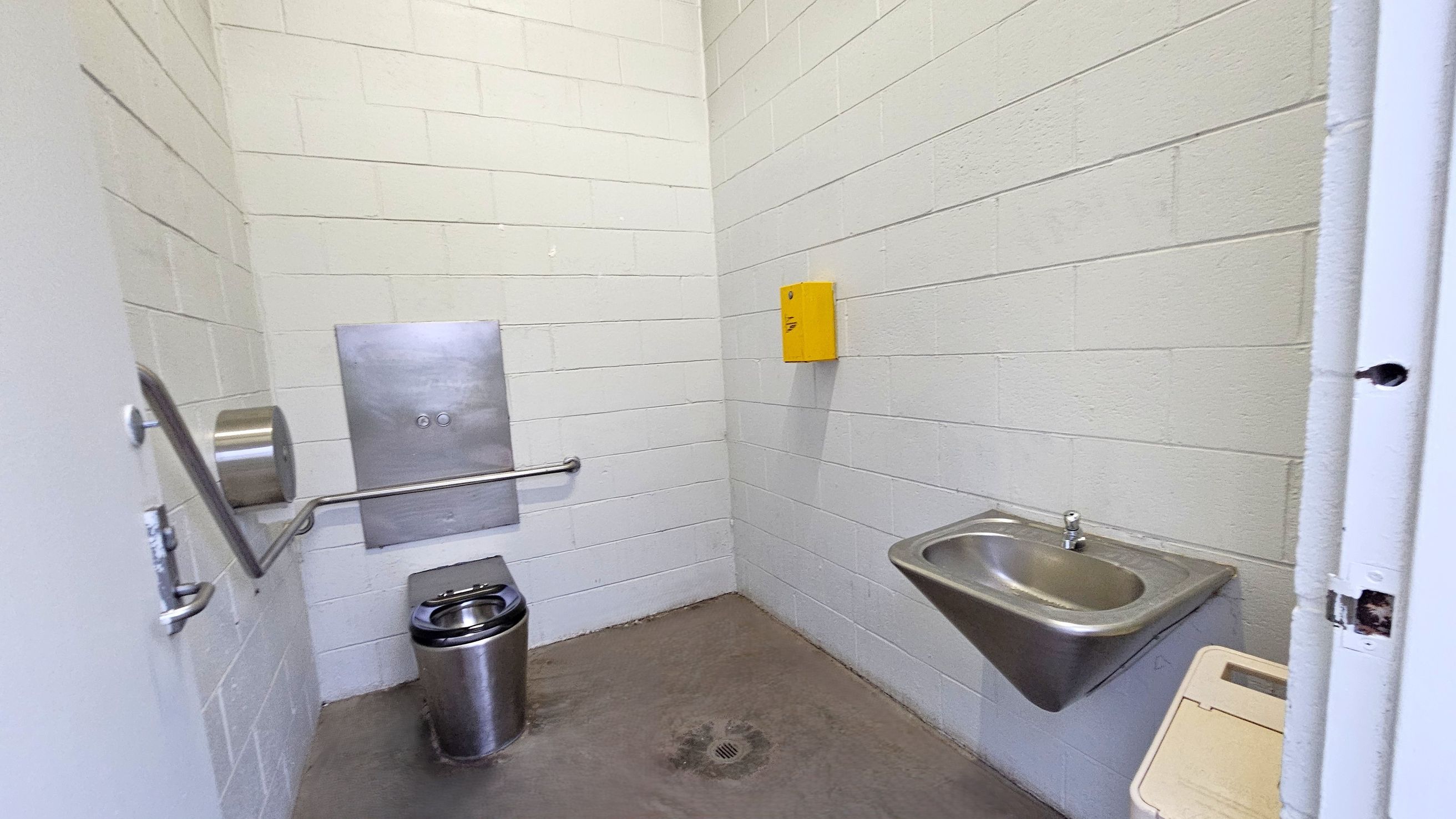 A small, stark bathroom with white cinder block walls. On the left, a stainless steel toilet with a metal toilet paper holder and a grab bar. In the centre, a metal panel is mounted on the wall. On the right, a stainless steel sink with a tap is attached to the wall, and a yellow box is mounted above it. The floor is concrete with a drain in the centre. The ceiling is plain and light-coloured.