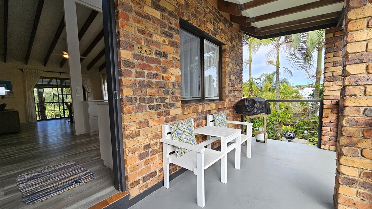 On the left, there is an interior space with wooden beams on the ceiling, a rug on the wooden floor, and a view of a living area with large windows. In the middle, there is a brick wall with a window and a small balcony area. On the right, the balcony has a white wooden bench with cushions, a barbecue grill, and potted plants. The floor of the balcony is concrete, and the ceiling has exposed wooden beams.