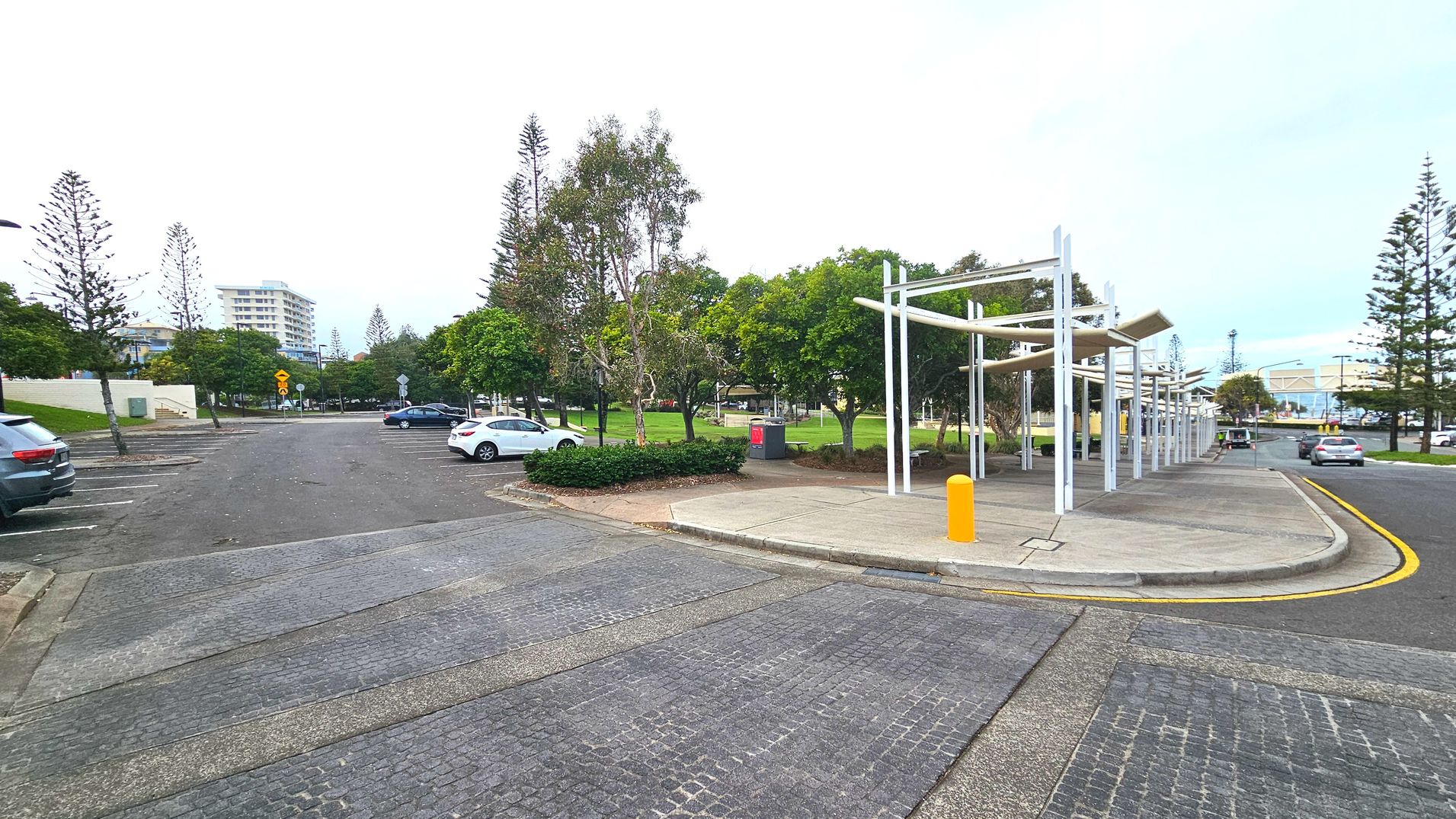 A paved area with a covered walkway featuring white metal beams and a yellow bollard. To the left, a car park with several parked cars is visible, bordered by trees and a grassy area. In the background, a multi-storey building rises above the trees. The ground transitions from cobblestone to smooth pavement. The space feels open with scattered trees and a clear view of the surrounding area.