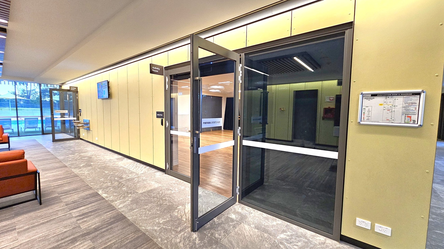 A modern interior with a glass door on the right leading to an activity room with wooden flooring. The wall is light-coloured with a mounted evacuation sign and diagram. On the left, there are red cushioned chairs and a large window showing an outdoor area with seating. The floor is tiled in a grey pattern, and the ceiling has recessed lighting. A digital screen is mounted on the wall near the door.