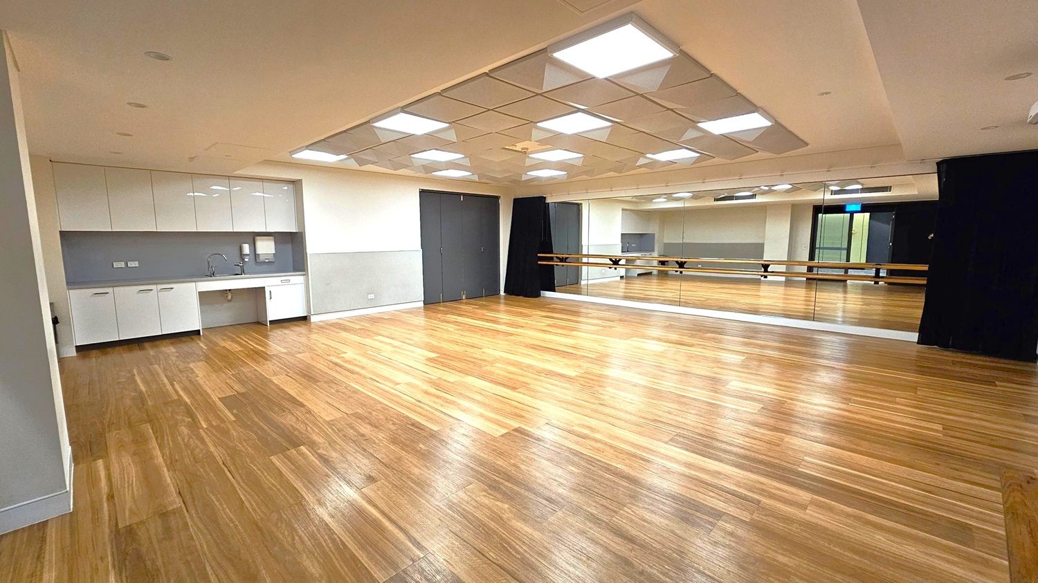 A spacious room with a wooden floor and a ceiling featuring recessed lighting. On the left, there is a kitchenette with white cabinets, a sink, and a paper towel dispenser. The right side has a large mirror covering the wall, with a wooden ballet barre running across it. The back wall has dark double doors and a black curtain. The room is well-lit and appears to be a dance or rehearsal studio.