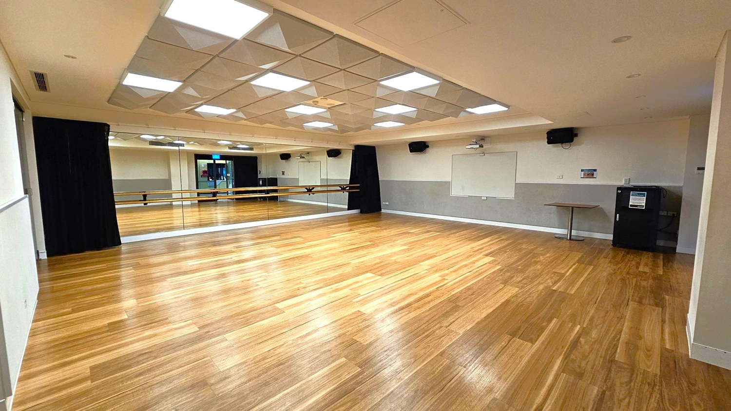 A spacious room with a wooden floor and a mirrored wall on the left. The ceiling features geometric light panels. On the right, there is a whiteboard, a small table, and a black cabinet. The room is equipped with speakers and a projector. The mirrored wall reflects the room, showing a door and additional lighting.