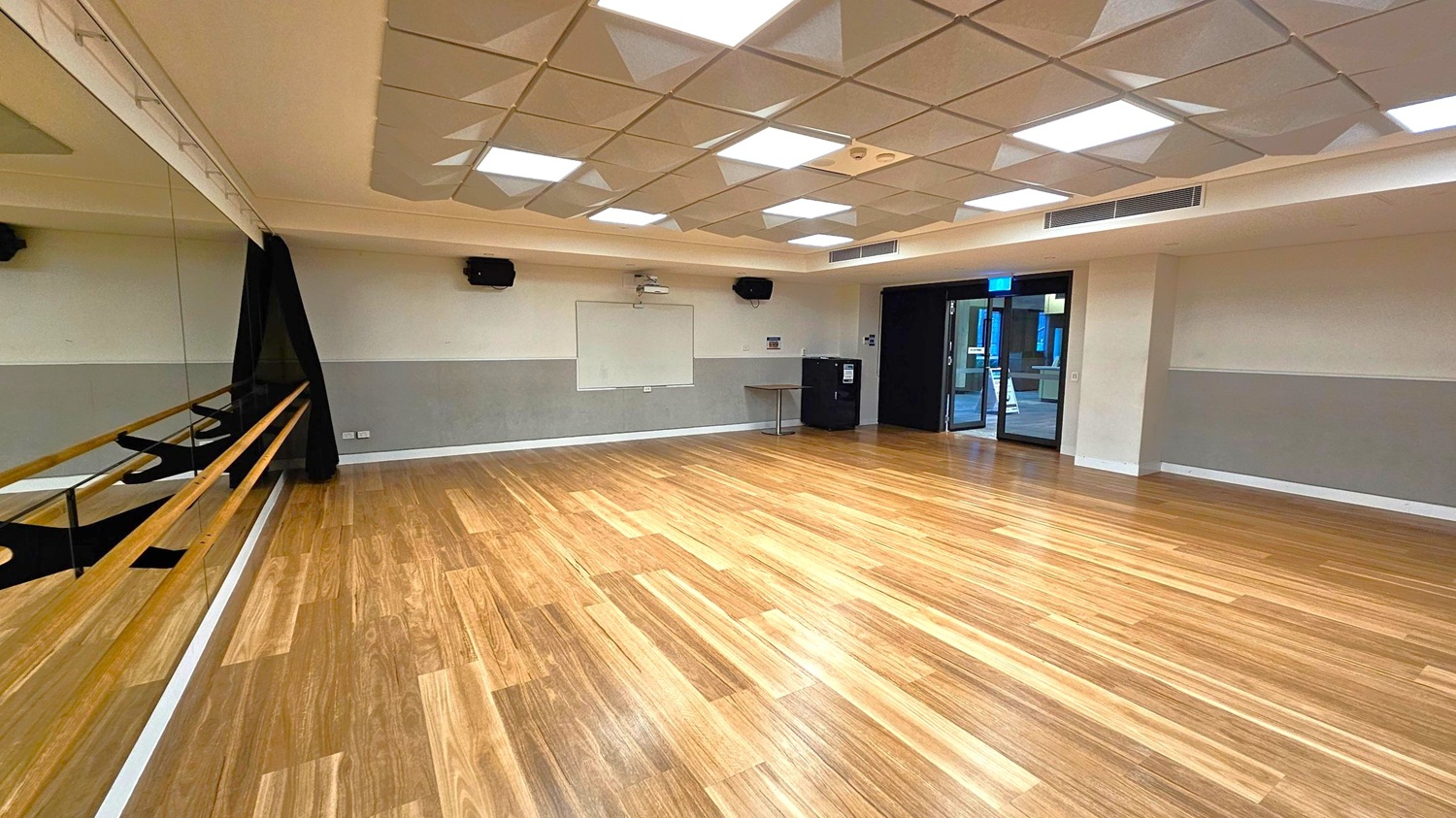 A spacious room with a wooden floor and a mirrored wall on the left, featuring wooden ballet barres. The ceiling has a geometric pattern with recessed lighting. On the far wall, there's a whiteboard flanked by speakers. A small table and a black cabinet are near the entrance, which has glass doors leading to another area. The walls are painted in two tones, with a darker shade on the lower half.