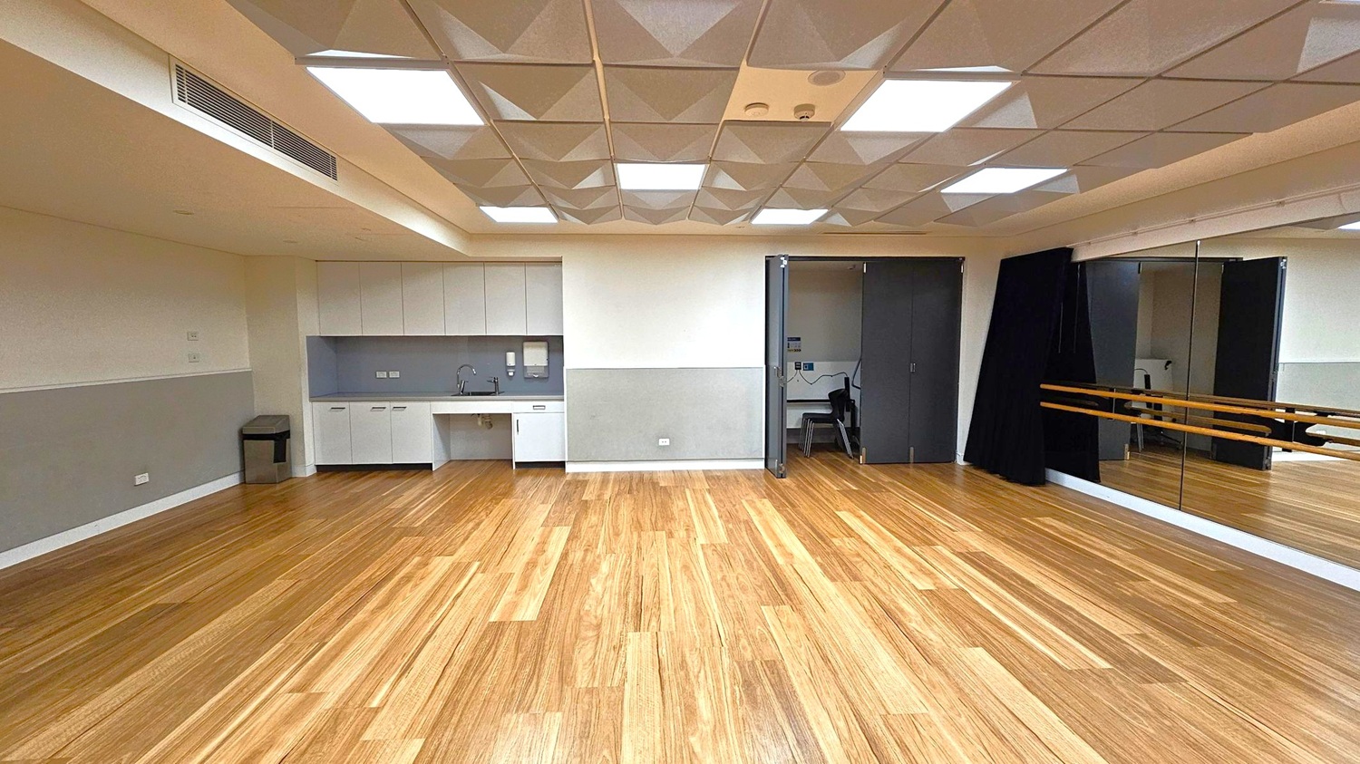 A spacious room with a wooden floor and a ceiling featuring geometric patterns and recessed lighting. On the left, there is a kitchenette with white cabinets, a sink, and a small bin. In the centre, a doorway leads to a smaller room with chairs. On the right, a large mirror covers the wall, with wooden ballet barres attached. The walls are painted in a light colour with a darker lower section.