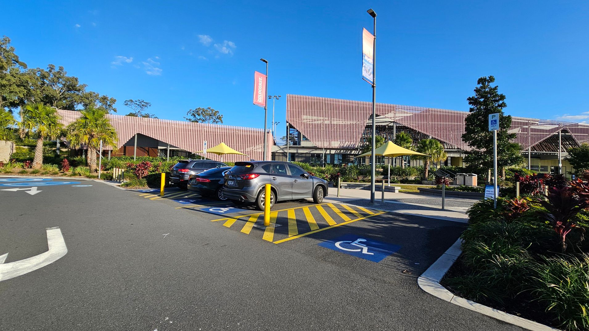 A car park with designated disabled parking spaces marked in blue and yellow is in the foreground. Three cars are parked in these spaces. The background features a modern building with a slatted facade and large windows. Palm trees and landscaped greenery are visible around the building. Two tall banners are mounted on poles, and there are yellow shade structures near the entrance. The sky is clear and blue.