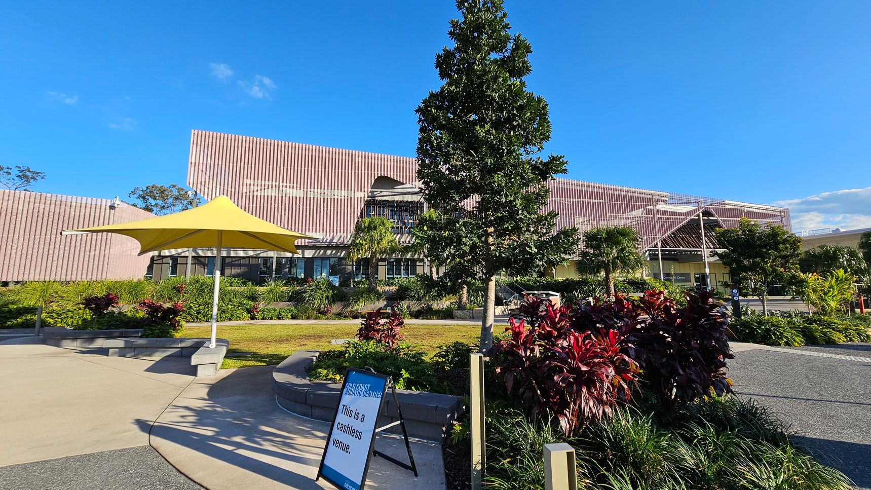 A modern building with a textured facade is in the background, surrounded by lush greenery and various plants. In the foreground, a yellow umbrella provides shade over a small seating area. A sign reads "This is a cashless venue." A tree stands prominently in the centre, with a mix of red and green foliage around it. The ground is a combination of concrete and gravel paths, leading towards the building's entrance.