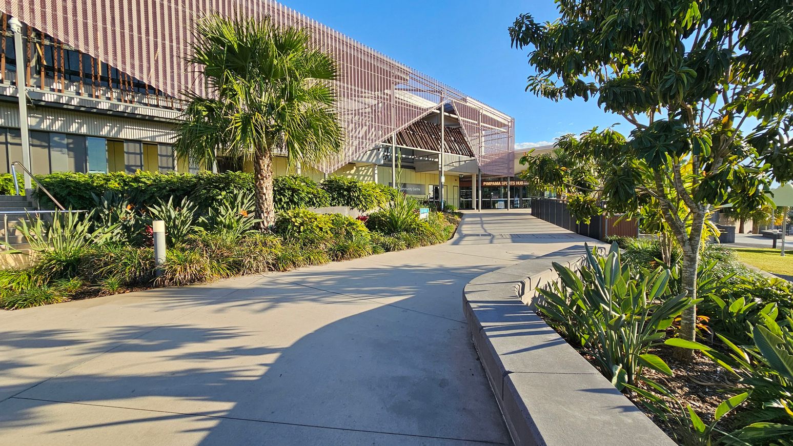 A modern building with a textured facade on the left, surrounded by lush greenery including palm trees and various plants. A wide concrete pathway curves towards the entrance, which is visible in the centre. On the right, more trees and plants line the path, with a low wall bordering the garden area. The scene is well-lit, suggesting a sunny day.