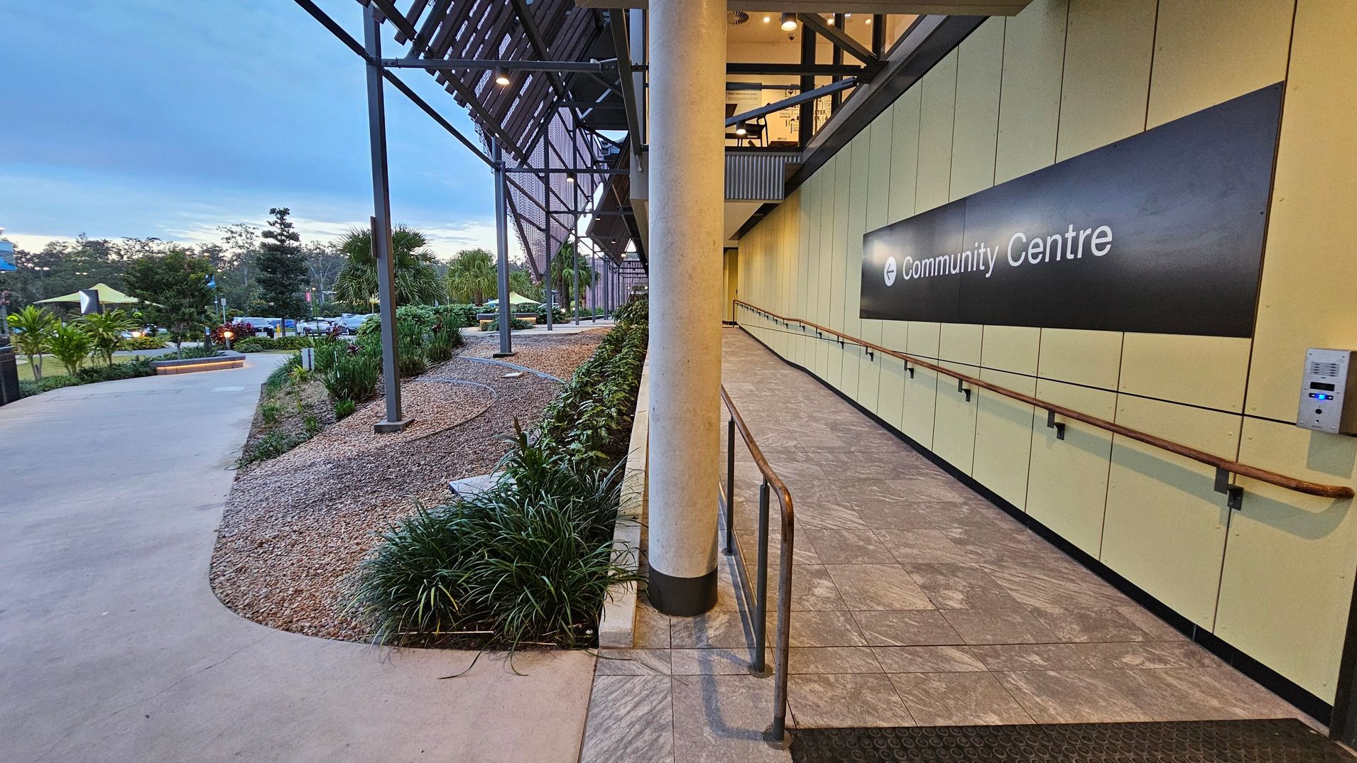 A pathway leads to a community centre on the right, with a sign on a light-coloured wall. The path is bordered by a metal railing and lined with small plants and gravel. On the left, a concrete walkway curves through a landscaped area with various shrubs and trees. The structure above has a metal framework with lights. The ceiling is visible with exposed beams and lighting fixtures.