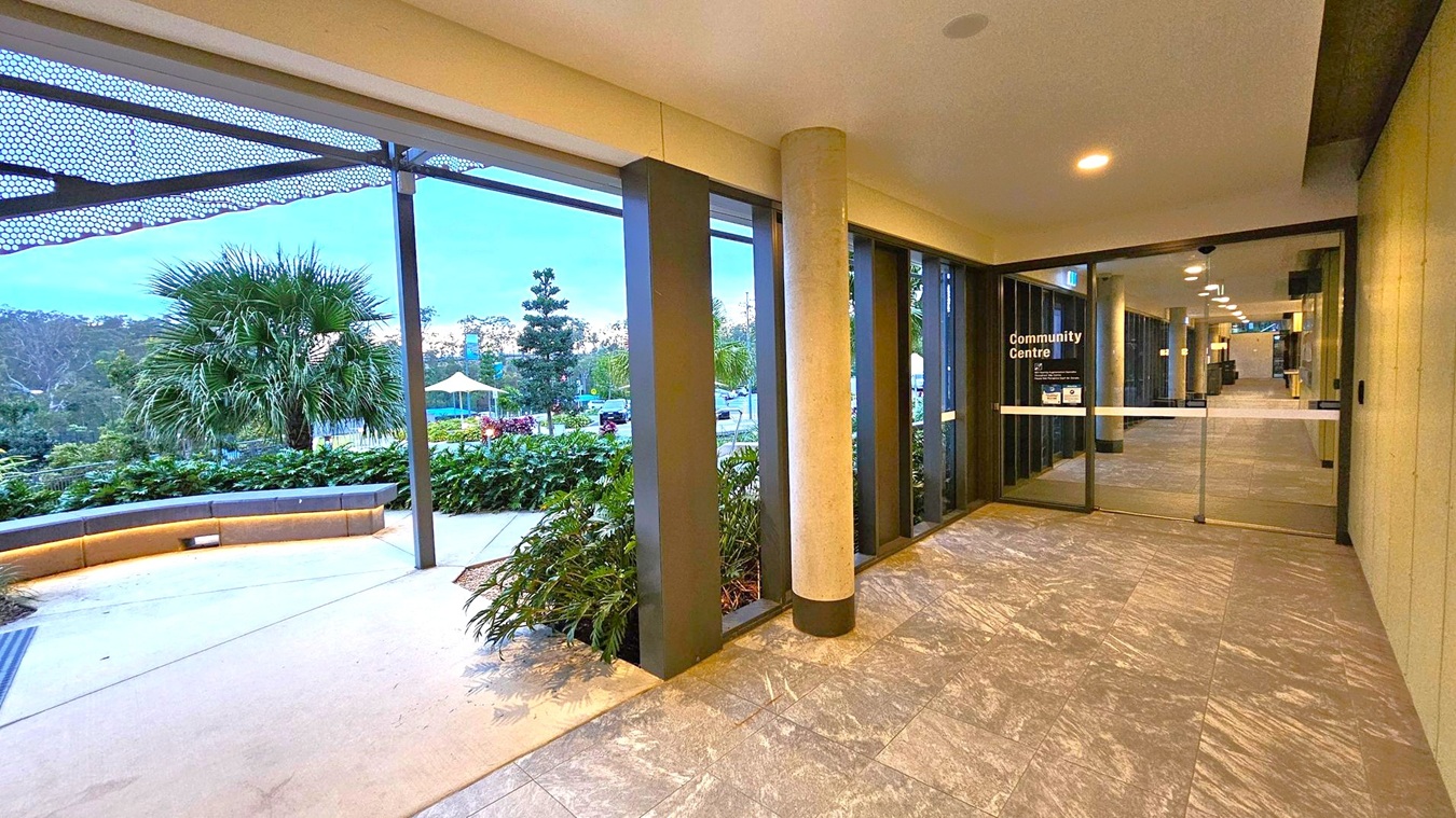 A corridor with a tiled floor leads to a glass door marked "Community Centre" on the right. The ceiling has recessed lighting. On the left, large windows reveal an outdoor area with a palm tree, greenery, and a curved bench. A metal canopy with a perforated design extends above the windows. In the background, there are more trees and a structure with a yellow canopy.
