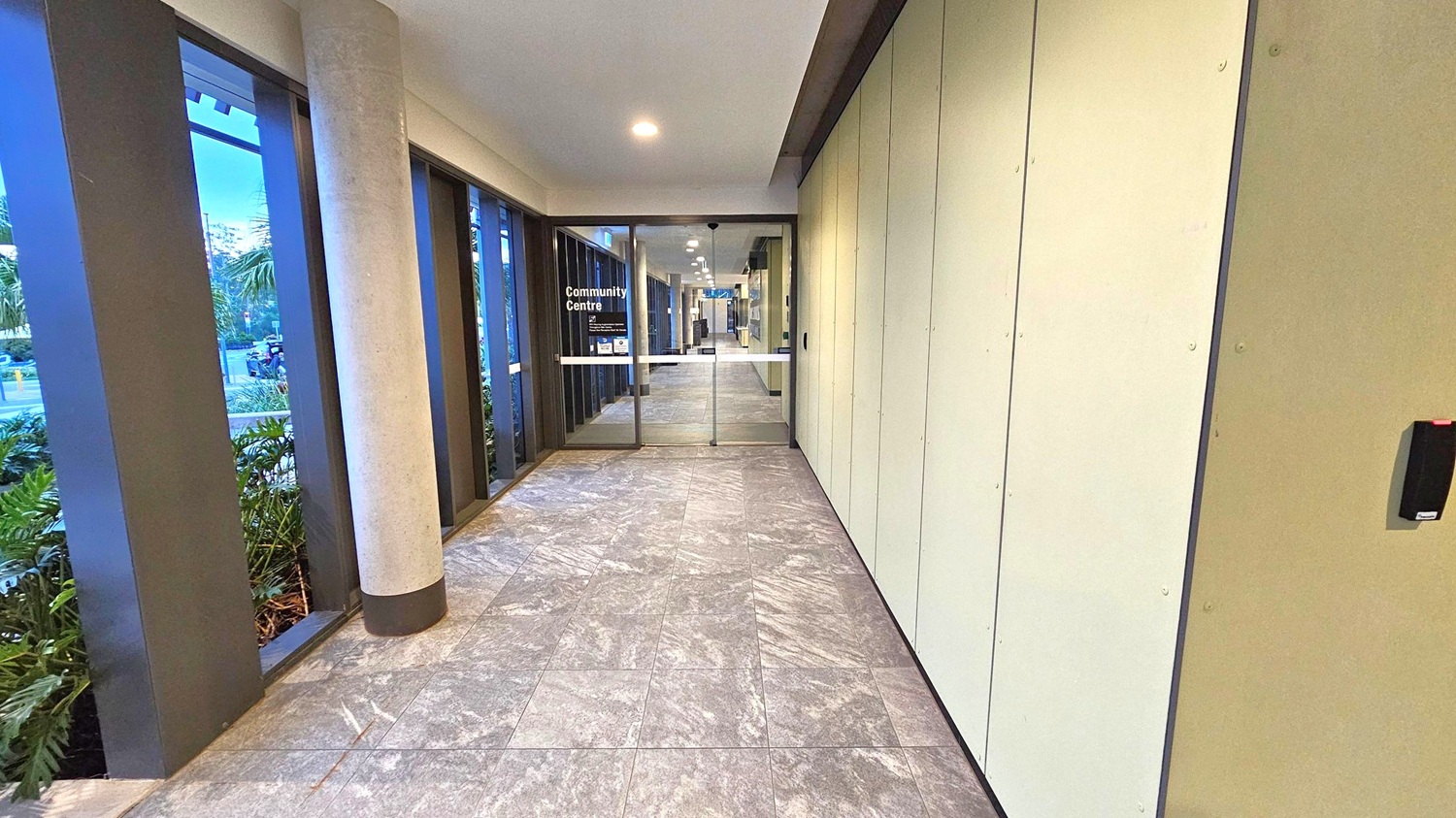 Corridor with a tiled floor in shades of grey and purple. On the left, large windows with vertical black frames and a concrete pillar reveal outdoor greenery. On the right, a plain beige wall with a black card reader. At the end, glass doors with "Community Centre" written on them, leading to a well-lit interior space. The ceiling is white with recessed lighting.