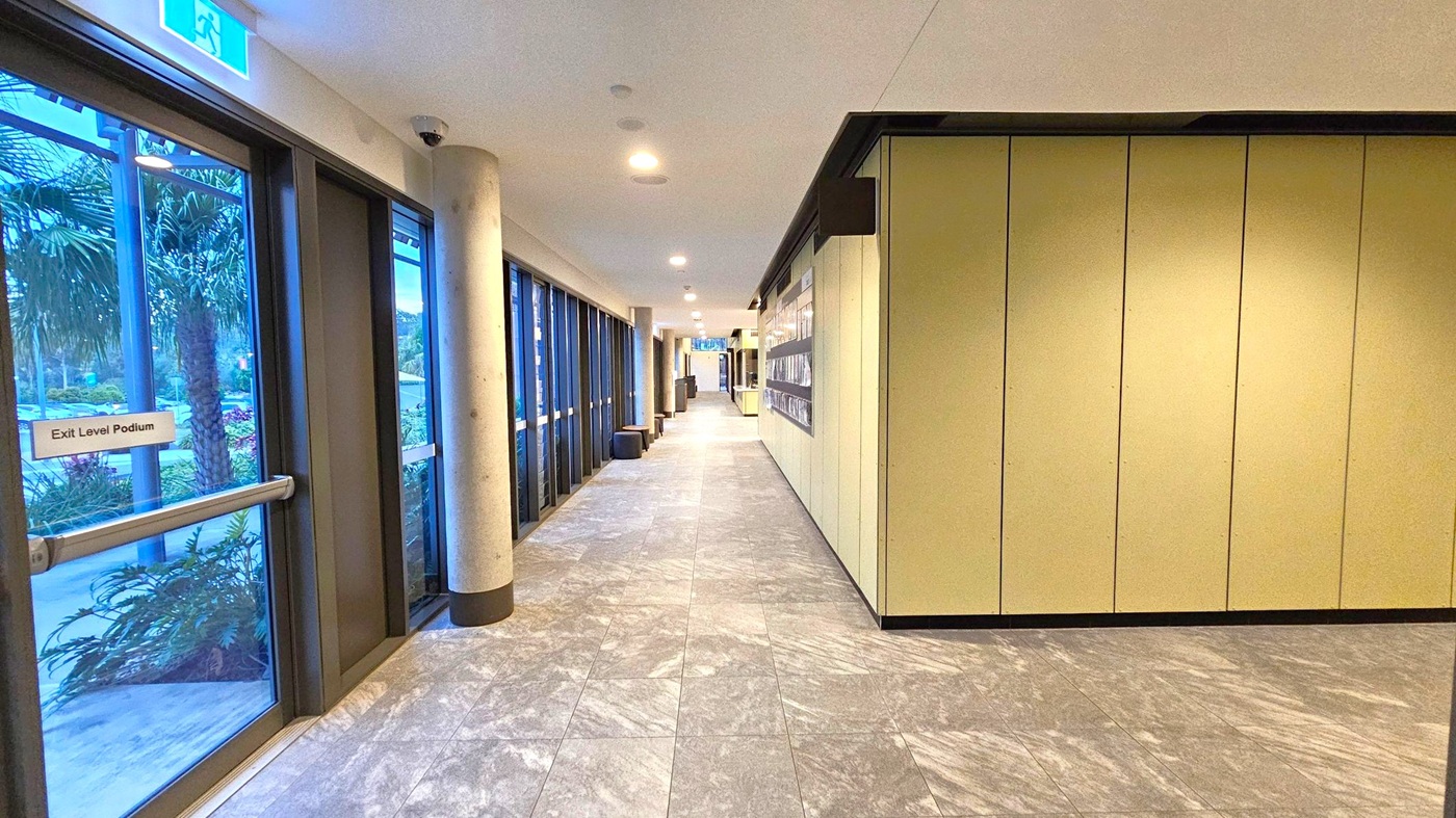 A corridor with a tiled floor and a ceiling with recessed lights. On the left, there are large glass doors with a sign reading "Exit Level Podium," offering a view of palm trees and greenery outside. On the right, there is a wall with vertical panels and a display case. A cylindrical column is visible near the glass doors. The corridor extends into the distance, with more doors and walls visible further down.