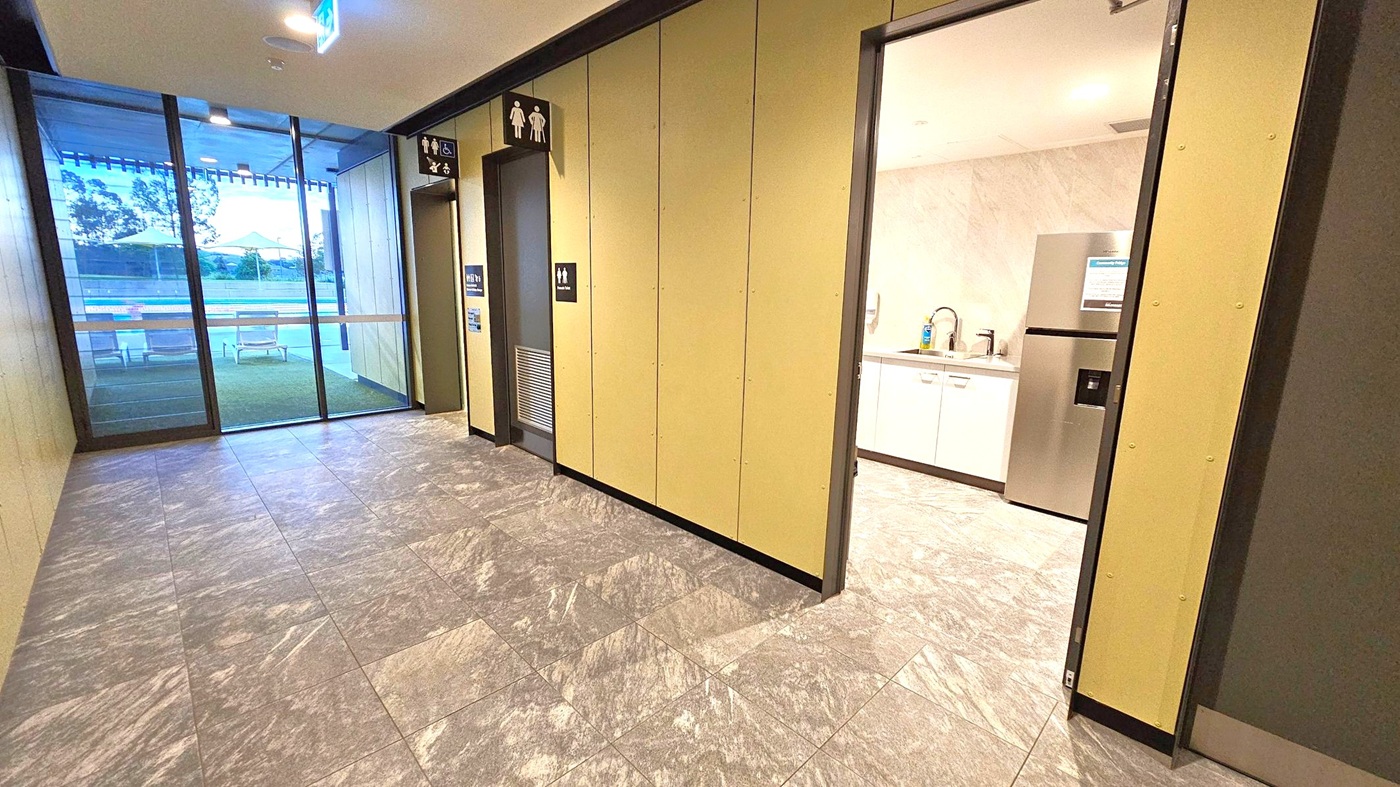 A hallway with grey marble flooring and beige walls. On the right, an open door reveals a kitchenette with a sink, white cabinets, and a stainless steel fridge. In the centre, a door with male and female restroom signs. On the left, large glass windows show an outdoor area with chairs and yellow umbrellas. The ceiling is plain with recessed lighting and an exit sign.
