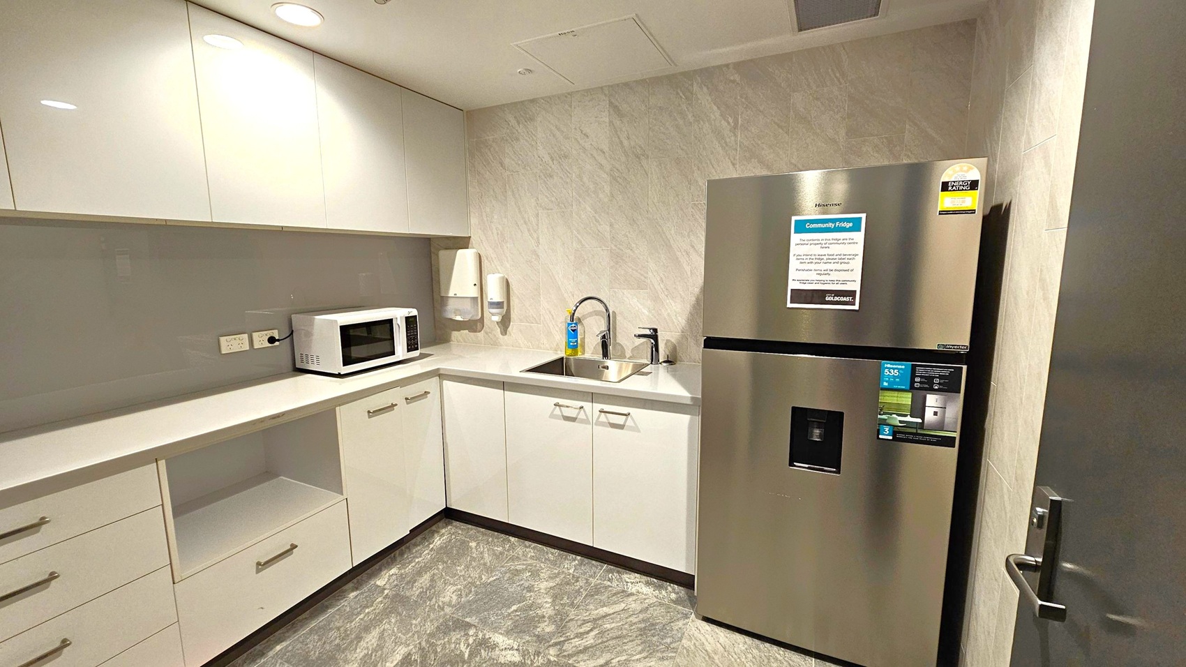 A compact kitchen with light-coloured cabinets and a grey marble-patterned floor. On the left, there are upper cabinets and a countertop with a microwave. Below are drawers and an open shelf. In the centre, a sink with a tap is set into the counter, with a soap dispenser nearby. On the right, a stainless steel refrigerator with an energy rating sticker is positioned next to a door. The walls have a light, textured finish, and the ceiling is fitted with recessed lighting.